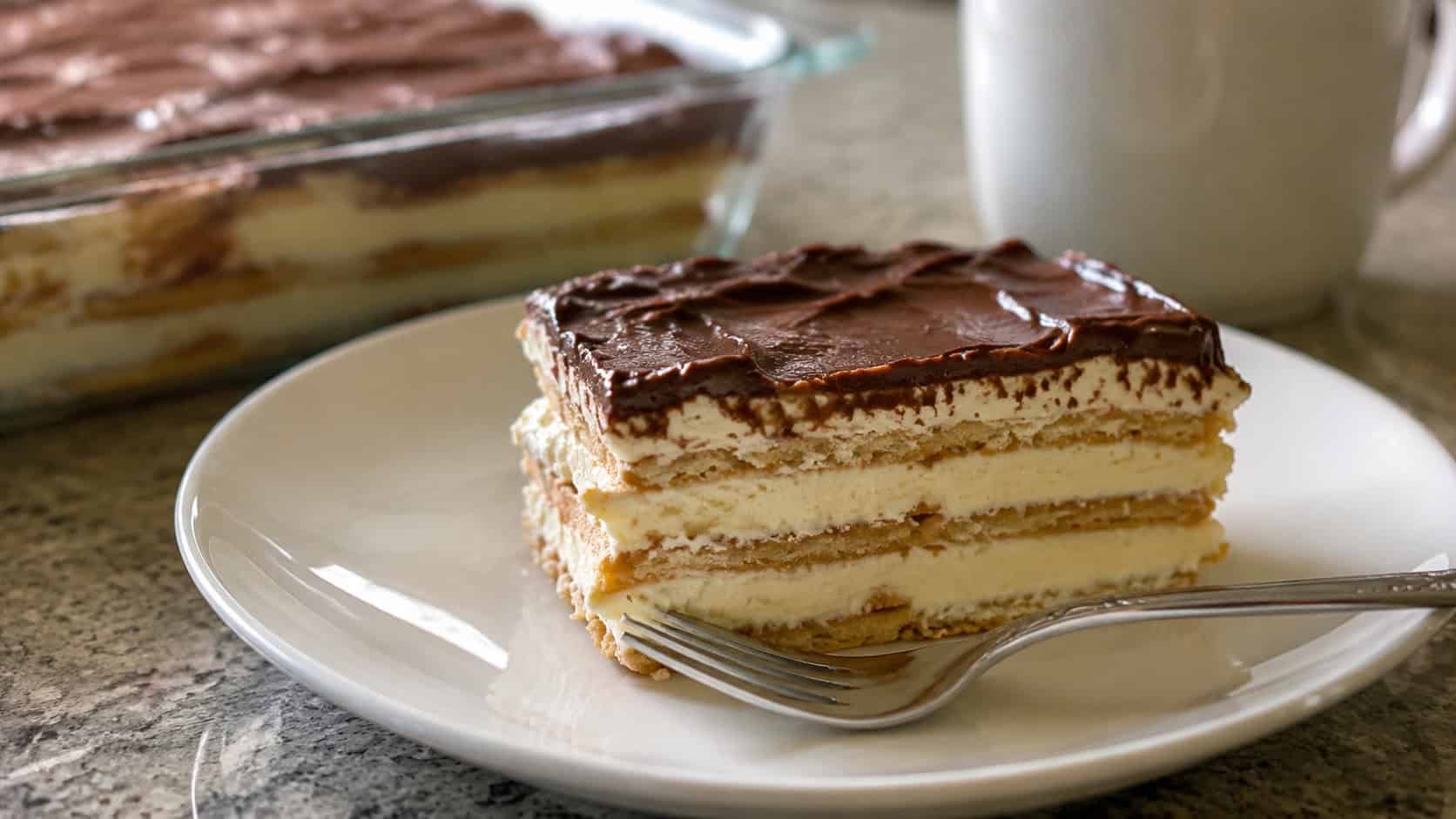 a slice of chocolate eclair cake with creamy layers and glossy chocolate top in a wide horizontal shot, on a white plate with a fork, soft natural light, shallow depth of field, landscape orientation