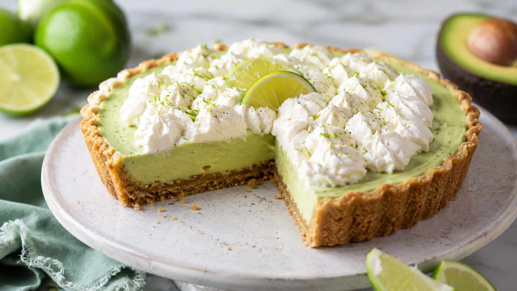 a pale green avocado lime tart topped with whipped cream and lime zest in a wide horizontal shot, on a ceramic stand with lime wedges, bright natural light, shallow depth of field, landscape orientation