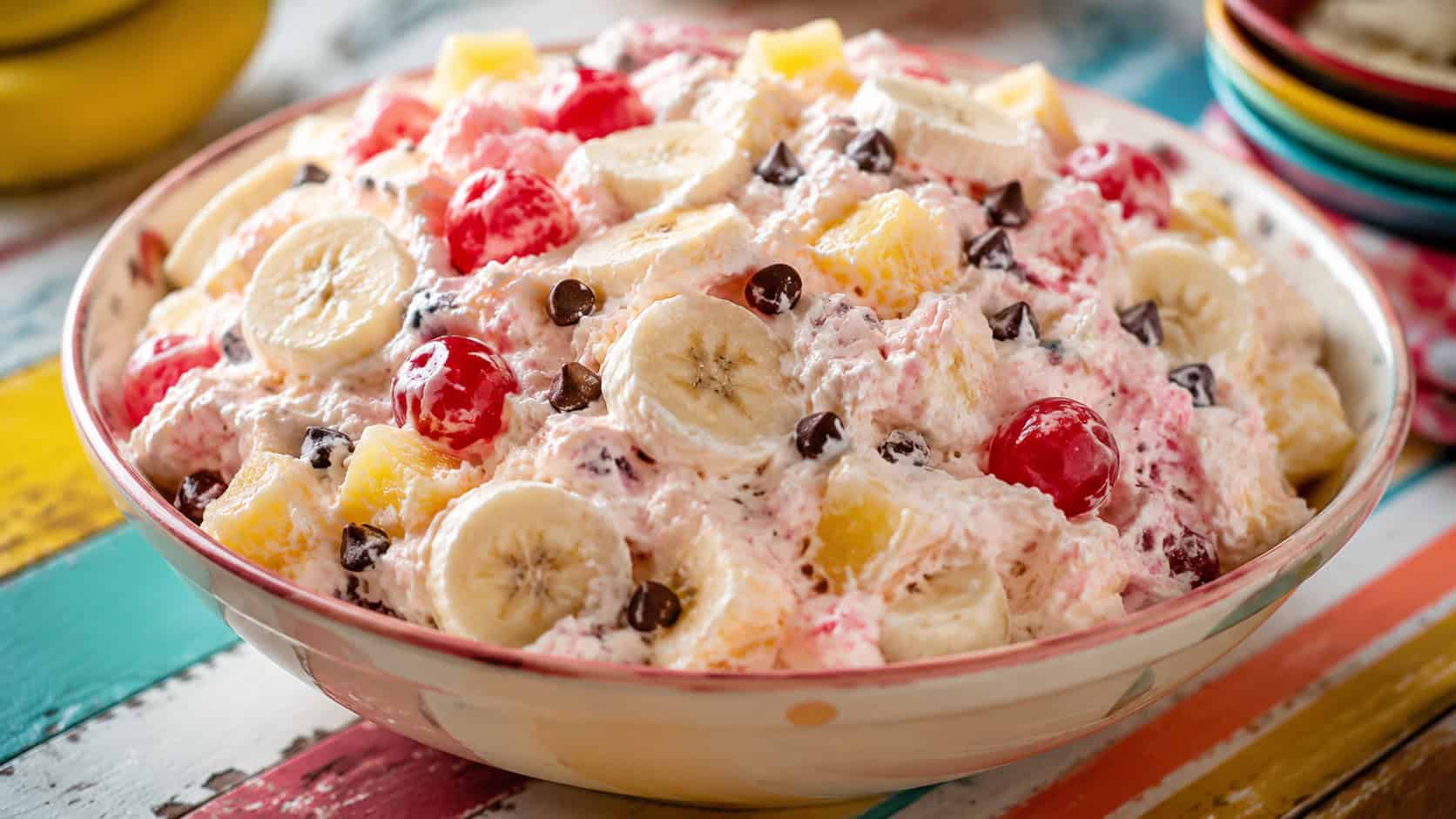 a big bowl of banana split fluff with bananas, pineapple, cherries, and chocolate chips in a wide horizontal shot, on a colorful table, bright natural light, shallow depth of field, landscape orientation