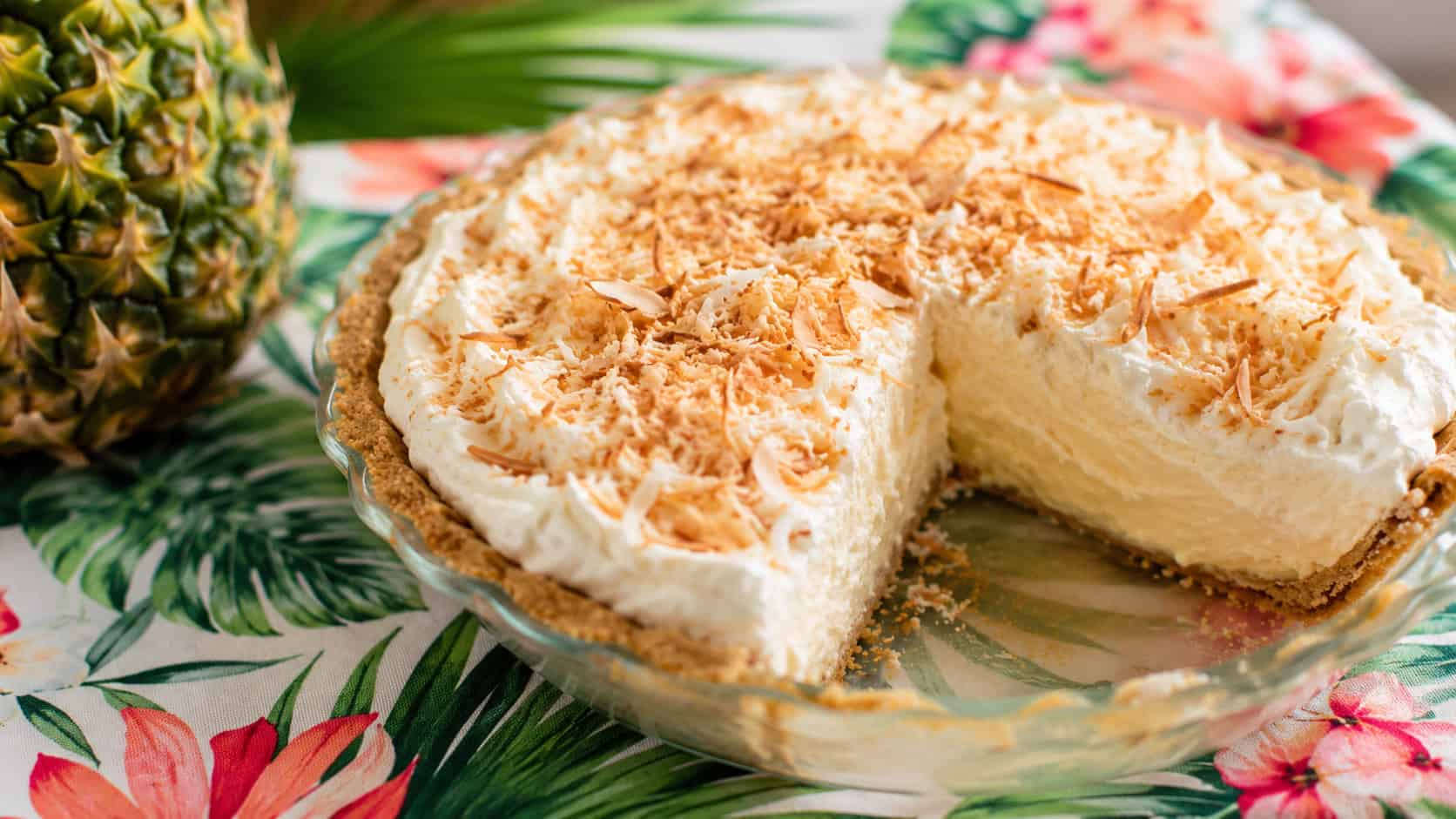 a creamy pina colada pie topped with toasted coconut in a wide horizontal shot, on a tropical-print cloth with a slice removed, bright natural light, shallow depth of field, landscape orientation