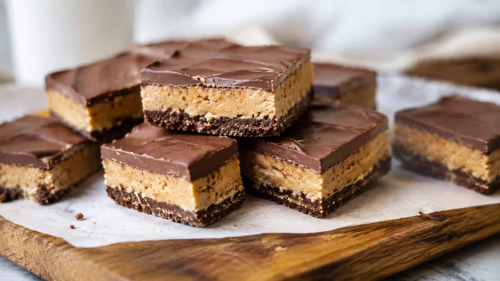 peanut butter chocolate bars cut into squares in a wide horizontal shot, stacked on parchment on a wooden board, soft natural light, shallow depth of field, landscape orientation
