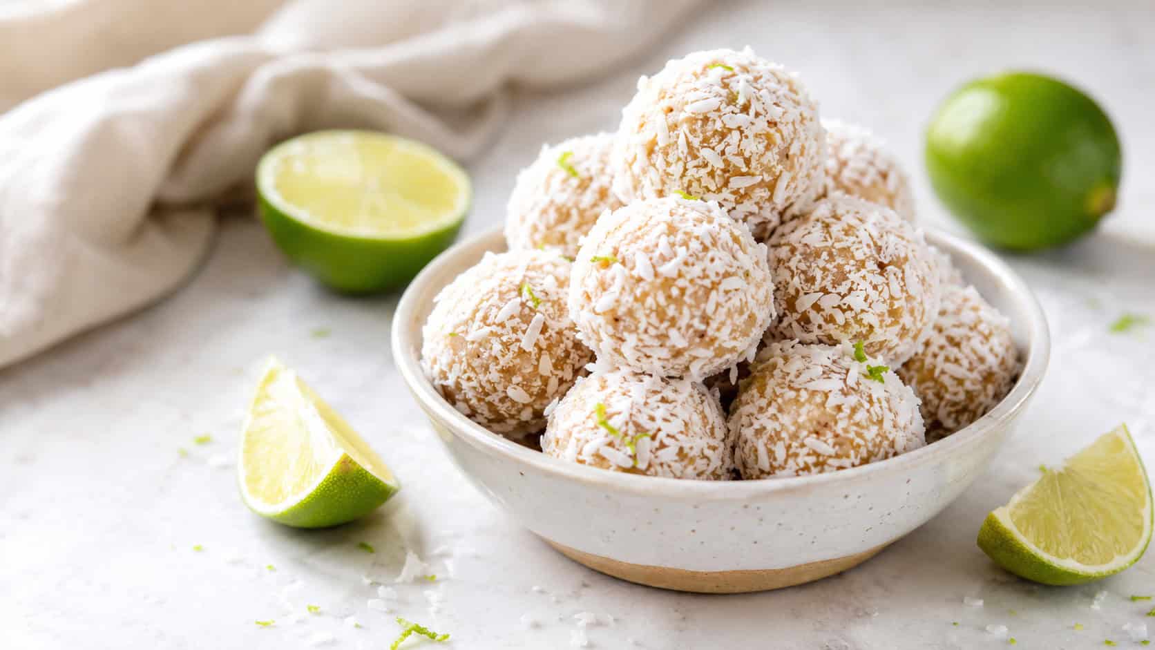 coconut lime energy bites rolled in shredded coconut in a wide horizontal shot, piled in a small bowl with lime wedges on a light counter, bright natural light, shallow depth of field, landscape orientation