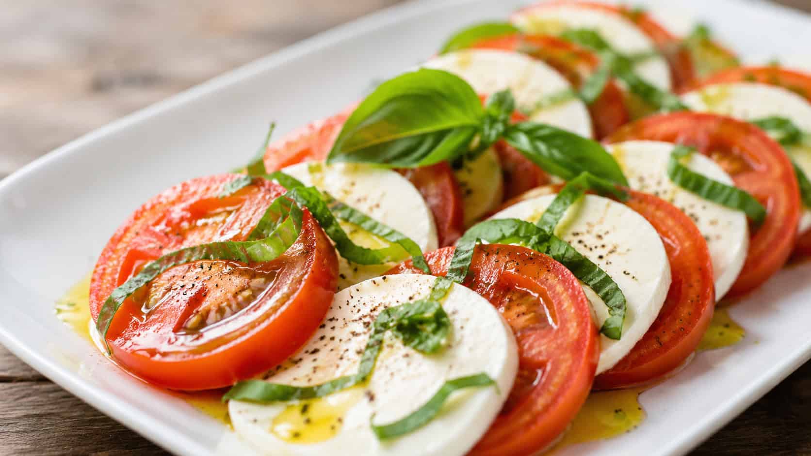 a caprese salad in a wide horizontal shot, overlapping red tomato and white mozzarella slices with green basil and olive oil on a white platter, natural light, shallow depth of field, landscape orientation