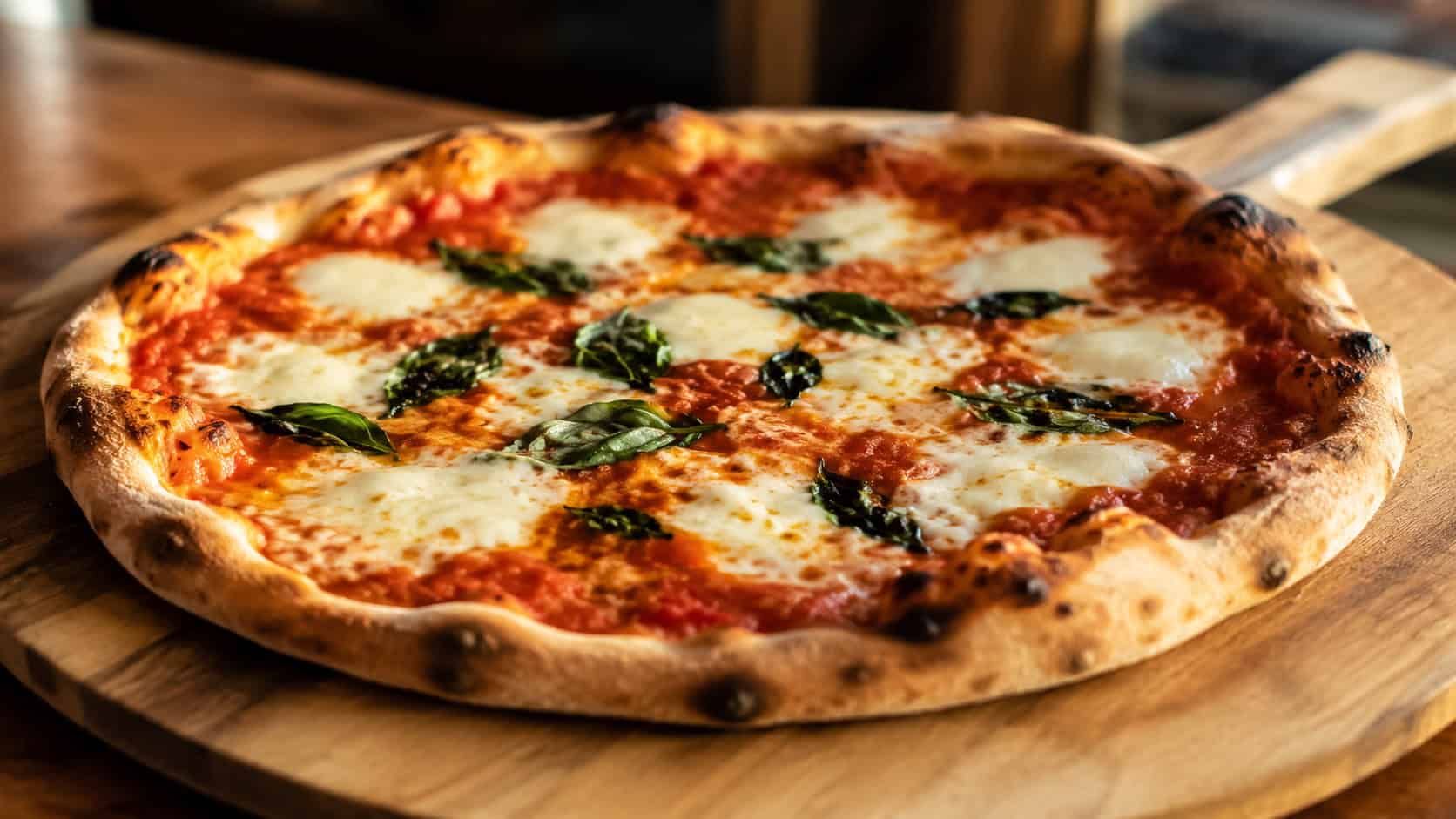 a margherita pizza in a wide horizontal shot, a whole pie with bubbly mozzarella, red sauce, and green basil on a wooden peel, warm natural light, shallow depth of field, landscape orientation