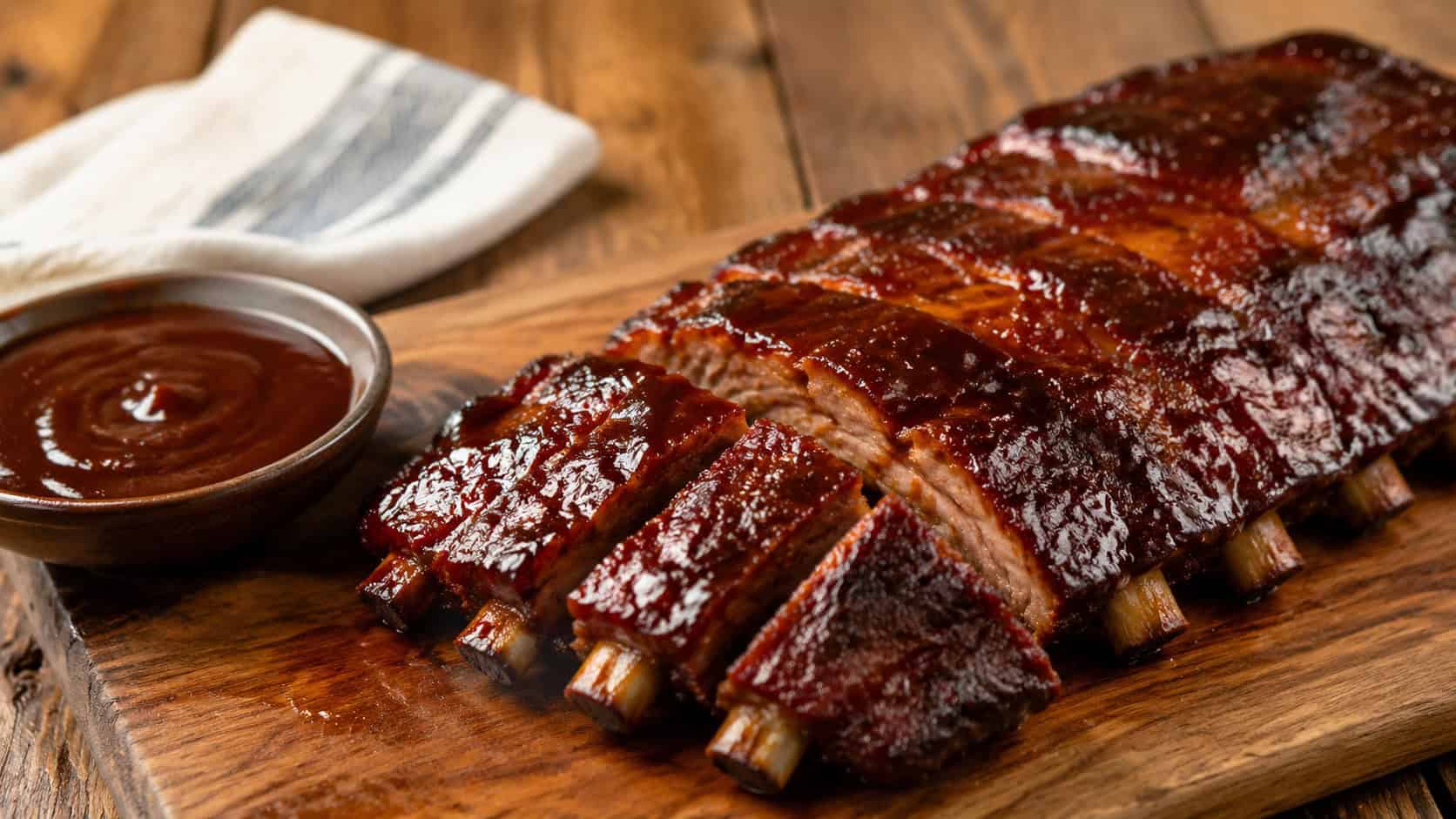 barbecue ribs on a wide wooden board shot horizontally, glossy caramelized ribs glistening with barbecue sauce, a small sauce bowl and napkin on a rustic surface, warm natural light, shallow depth of field, landscape orientation