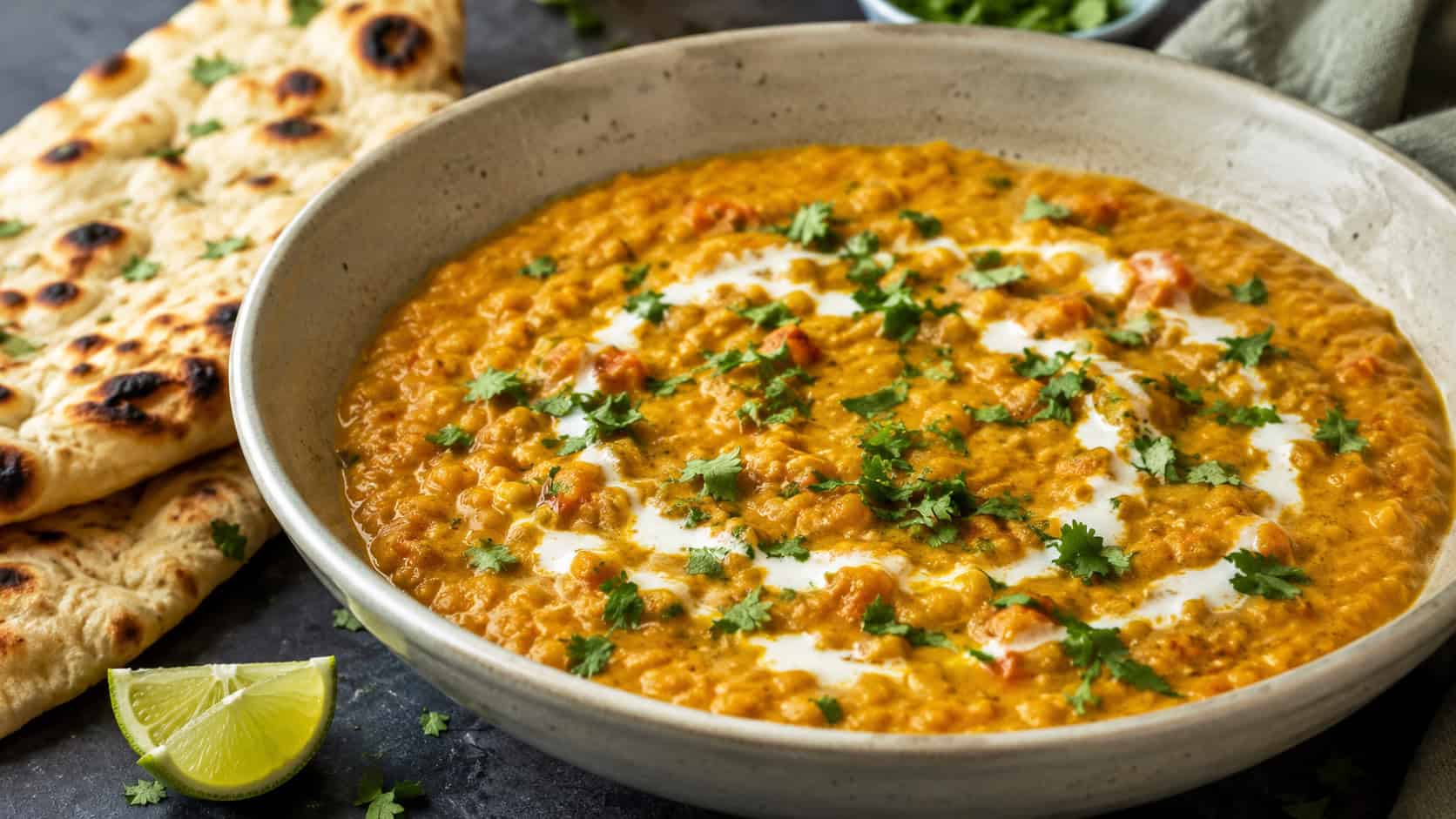 creamy red lentil curry in a wide bowl shot horizontally, thick golden curry topped with cilantro and a swirl of coconut milk, naan and a lime wedge on a dark surface, warm natural light, shallow depth of field, landscape orientation