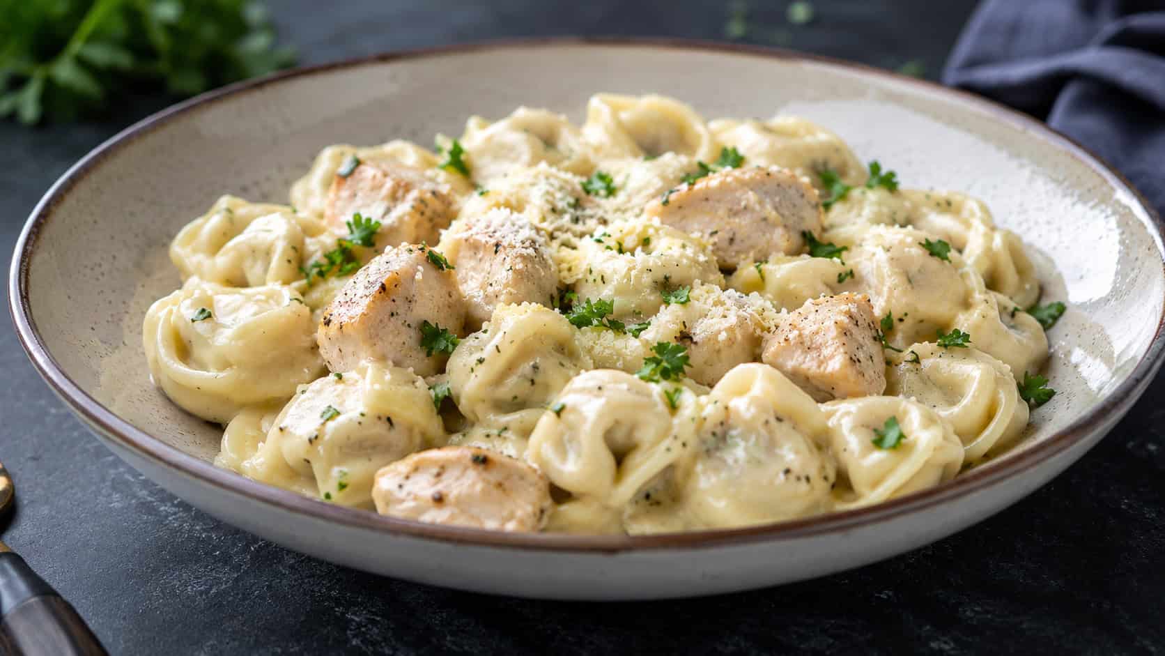 creamy chicken alfredo tortellini in a wide bowl shot horizontally, plump tortellini and chicken in a glossy white sauce dusted with parmesan and parsley, a dark elegant surface, soft natural light, shallow depth of field, landscape orientation