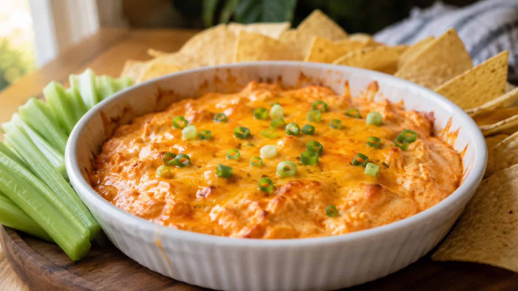 bubbling buffalo chicken dip in a wide dish shot horizontally, creamy orange dip topped with melted cheddar and green onions, celery sticks and tortilla chips fanned around it, warm natural light, shallow depth of field, landscape orientation
