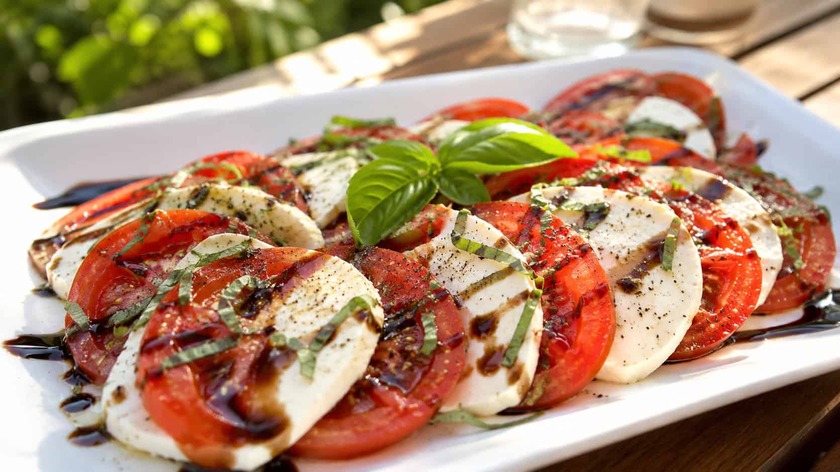 a caprese salad with overlapping tomato and mozzarella slices, fresh basil and balsamic drizzle on a white platter, on a sunny table, natural light, shallow depth of field, wide horizontal landscape composition