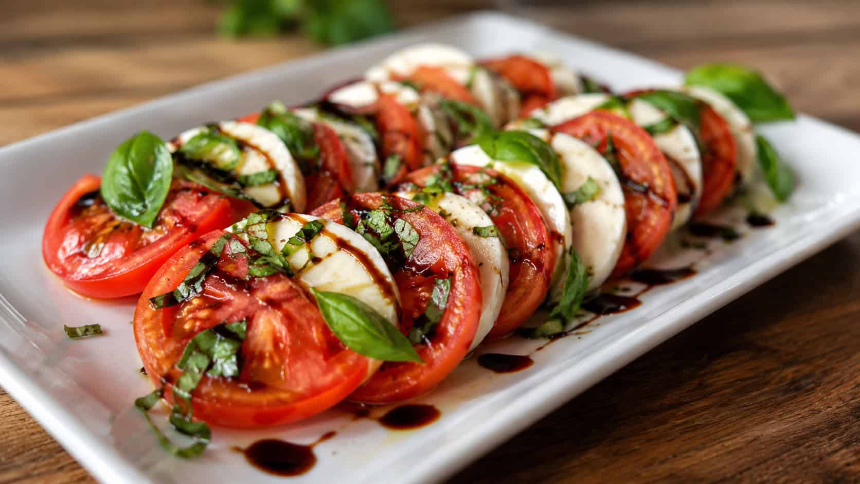 a caprese salad with layered tomato and mozzarella slices, fresh basil, and a balsamic drizzle in a wide horizontal shot, on a white platter over a wooden table, bright natural light, shallow depth of field, landscape orientation