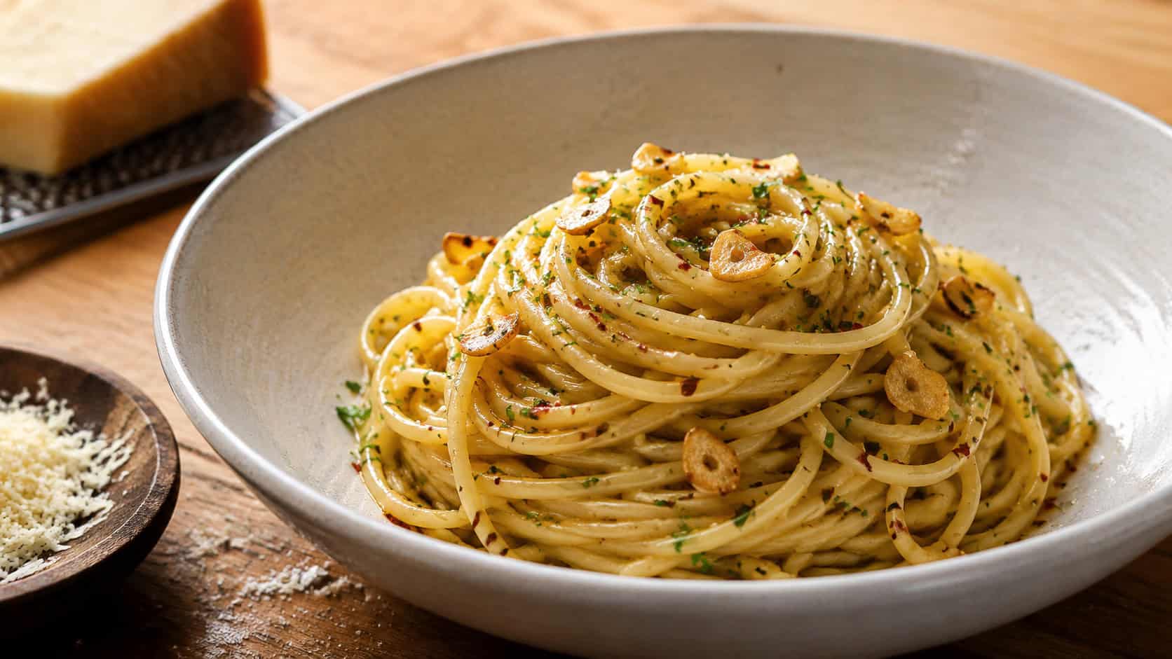 a twirl of glossy spaghetti tossed with golden garlic, chili flakes, and parsley in a wide horizontal shot, in a shallow bowl on a wooden table with grated parmesan nearby, warm natural light, shallow depth of field, landscape orientation