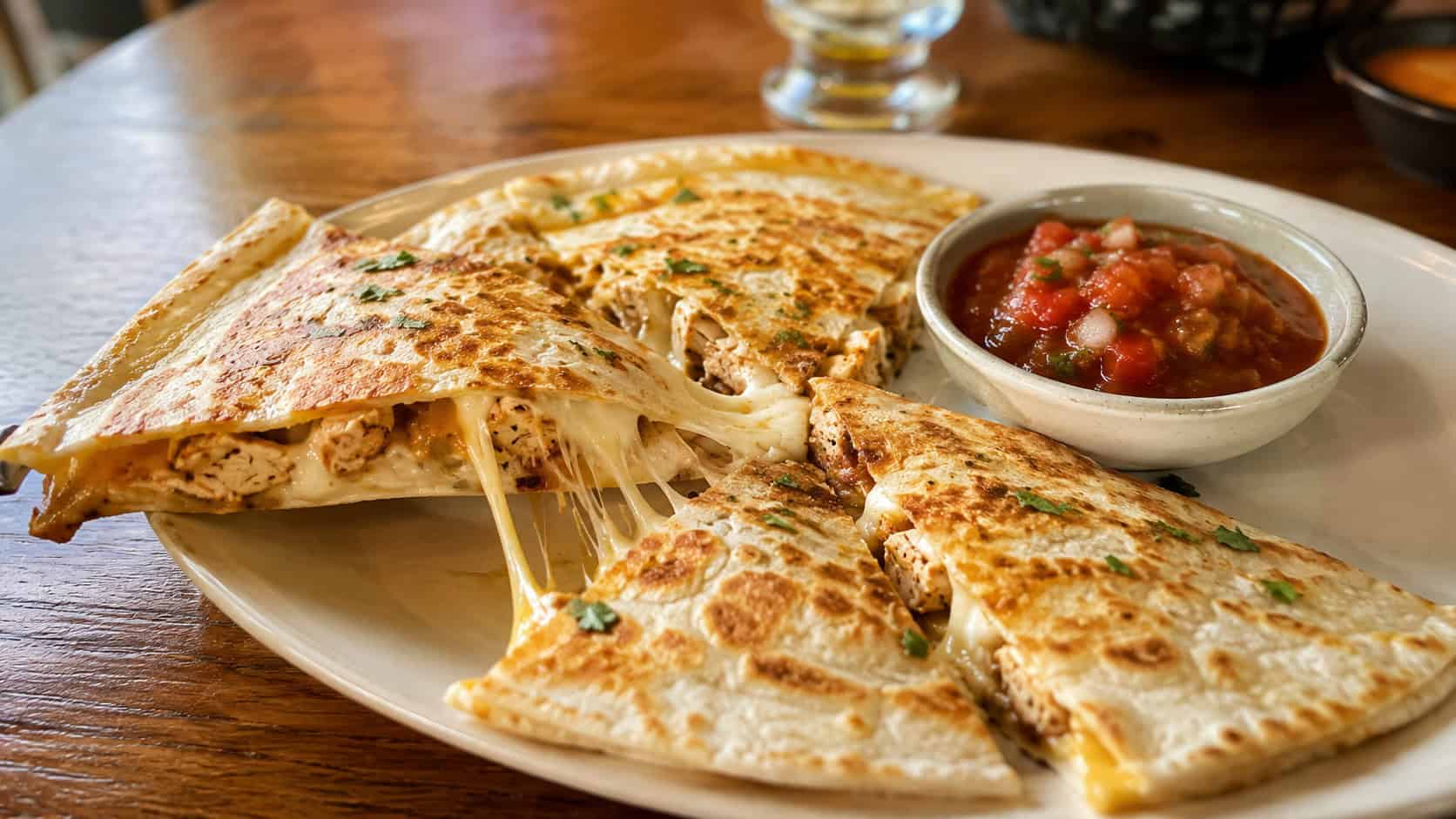 a golden chicken quesadilla cut into wedges with melty cheese stretching in a wide horizontal shot, on a plate with a bowl of salsa over a wooden table, warm natural light, shallow depth of field, landscape orientation