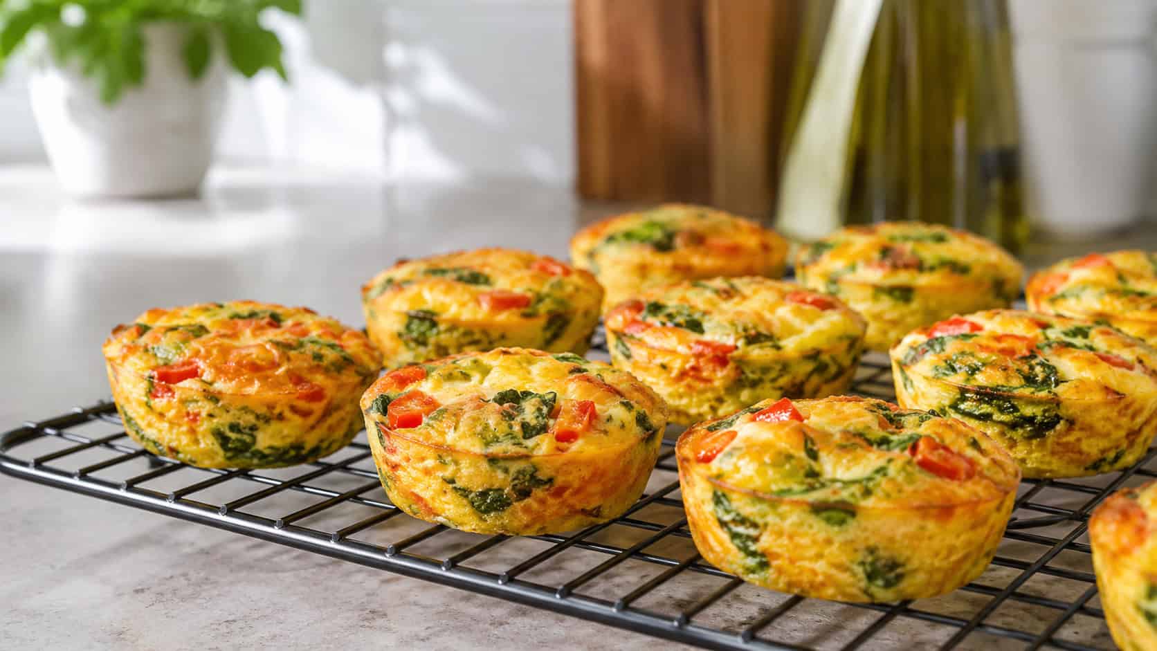 mini quiche egg muffins in a wide horizontal shot, golden cups flecked with red pepper and green spinach cooling on a wire rack beside a lunch box, bright kitchen light, shallow depth of field, landscape orientation