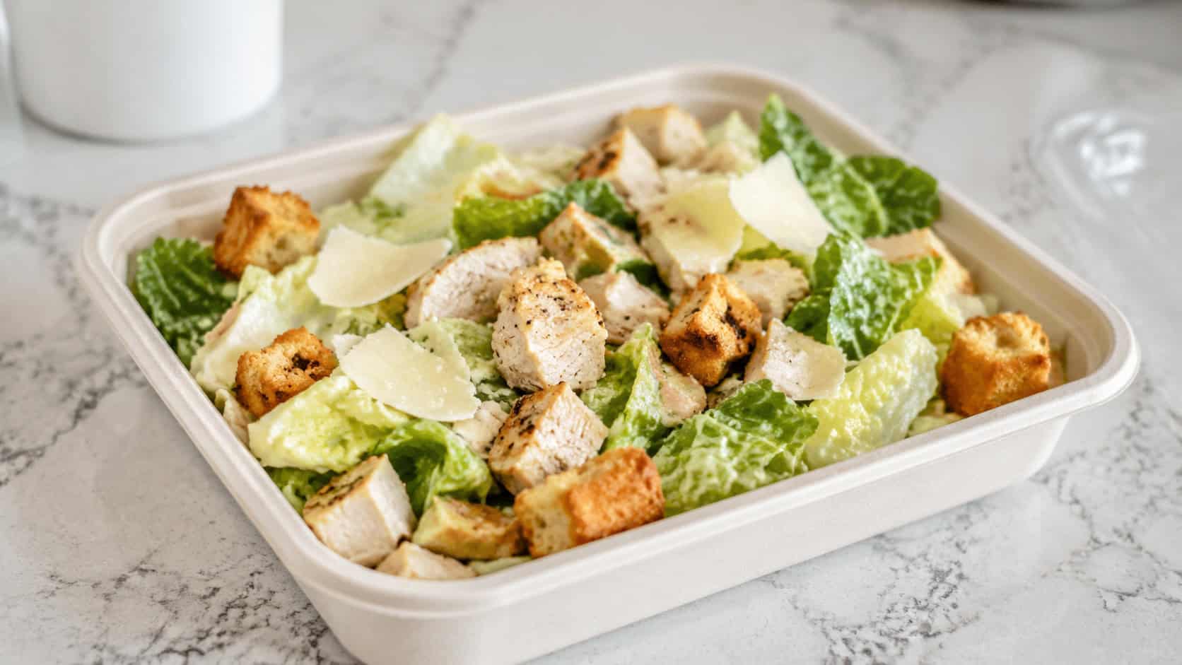 a chicken Caesar salad in a wide horizontal shot, crisp romaine topped with diced chicken, shaved parmesan, and golden croutons in a clear container, a small dressing cup beside it, bright natural light, shallow depth of field, landscape orientation