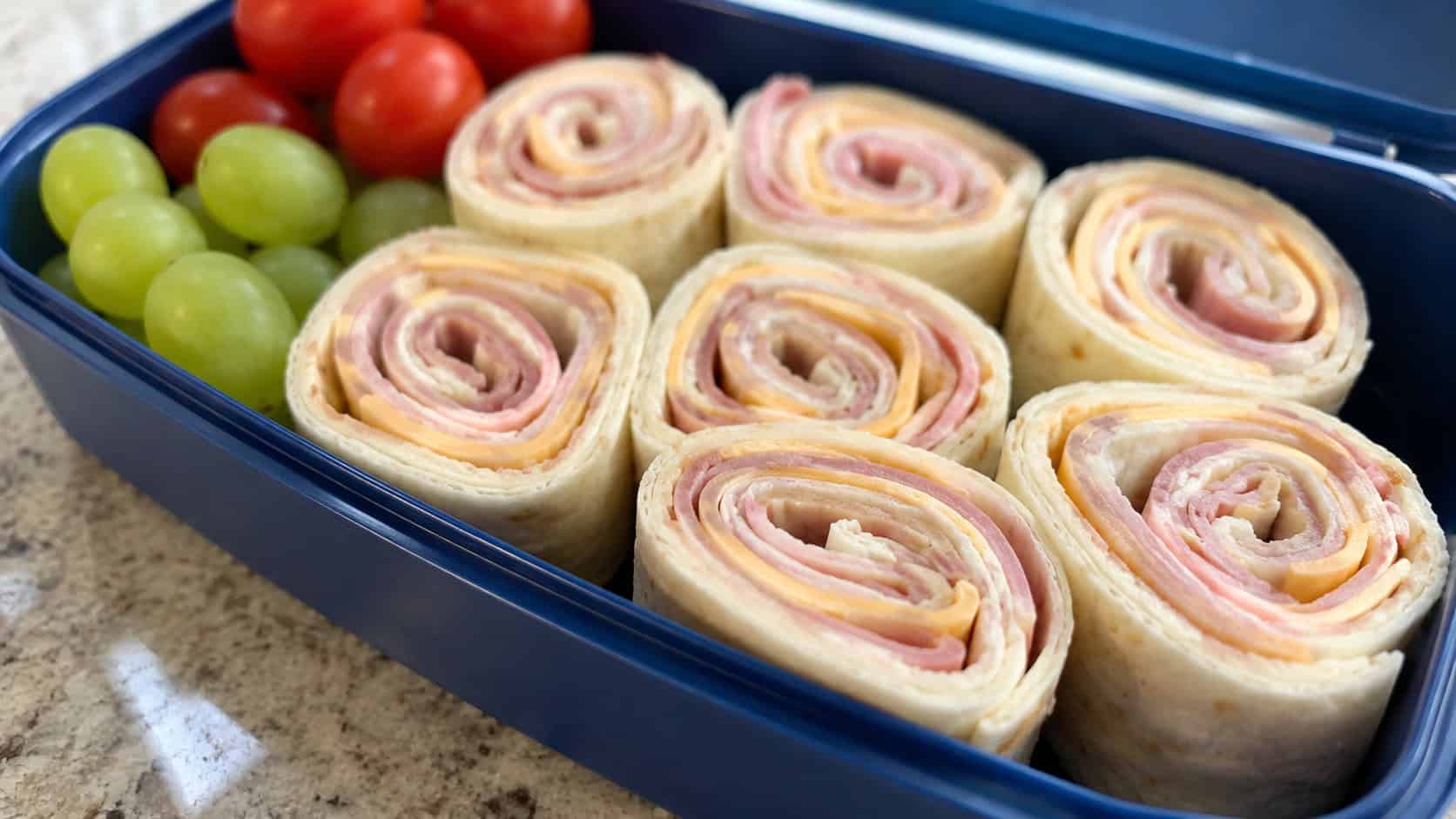 ham and cheese pinwheels in a wide horizontal shot, neat spirals showing ham, cheese, and green spinach arranged in a bento box with grapes, set on a bright counter, cheerful natural light, shallow depth of field, landscape orientation