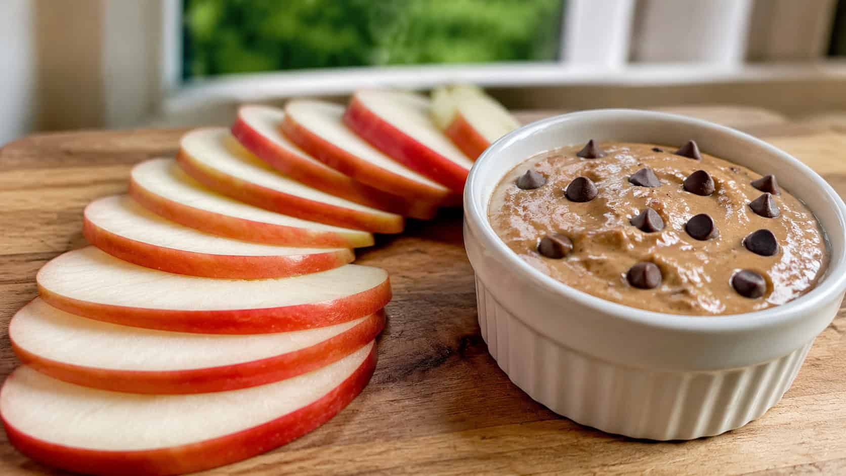 crisp apple slices fanned beside a small dish of almond butter in a wide horizontal shot, a sprinkle of chocolate chips on top, arranged in a bento lunch box, bright natural light, shallow depth of field, landscape orientation