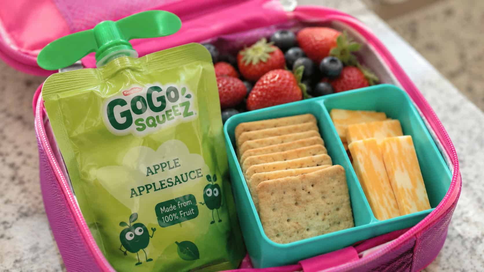 an applesauce pouch in a wide horizontal shot, the pouch beside gluten-free crackers and cheese slices in a colorful lunchbox, bright natural light, shallow depth of field, landscape orientation