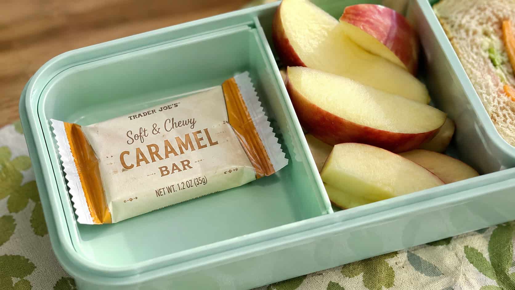 a wrapped caramel chewy bar in a wide horizontal shot, resting in a bento lunchbox next to fresh apple slices, bright natural light, shallow depth of field, landscape orientation