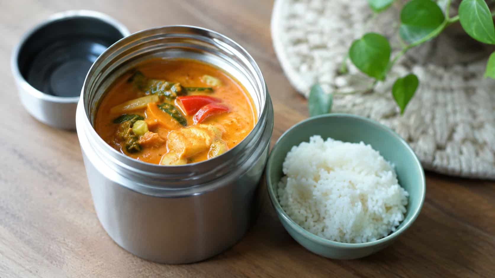 Thai red curry in a wide horizontal shot, a vibrant orange curry with vegetables in an open thermos beside a small bowl of rice, natural light, shallow depth of field, landscape orientation