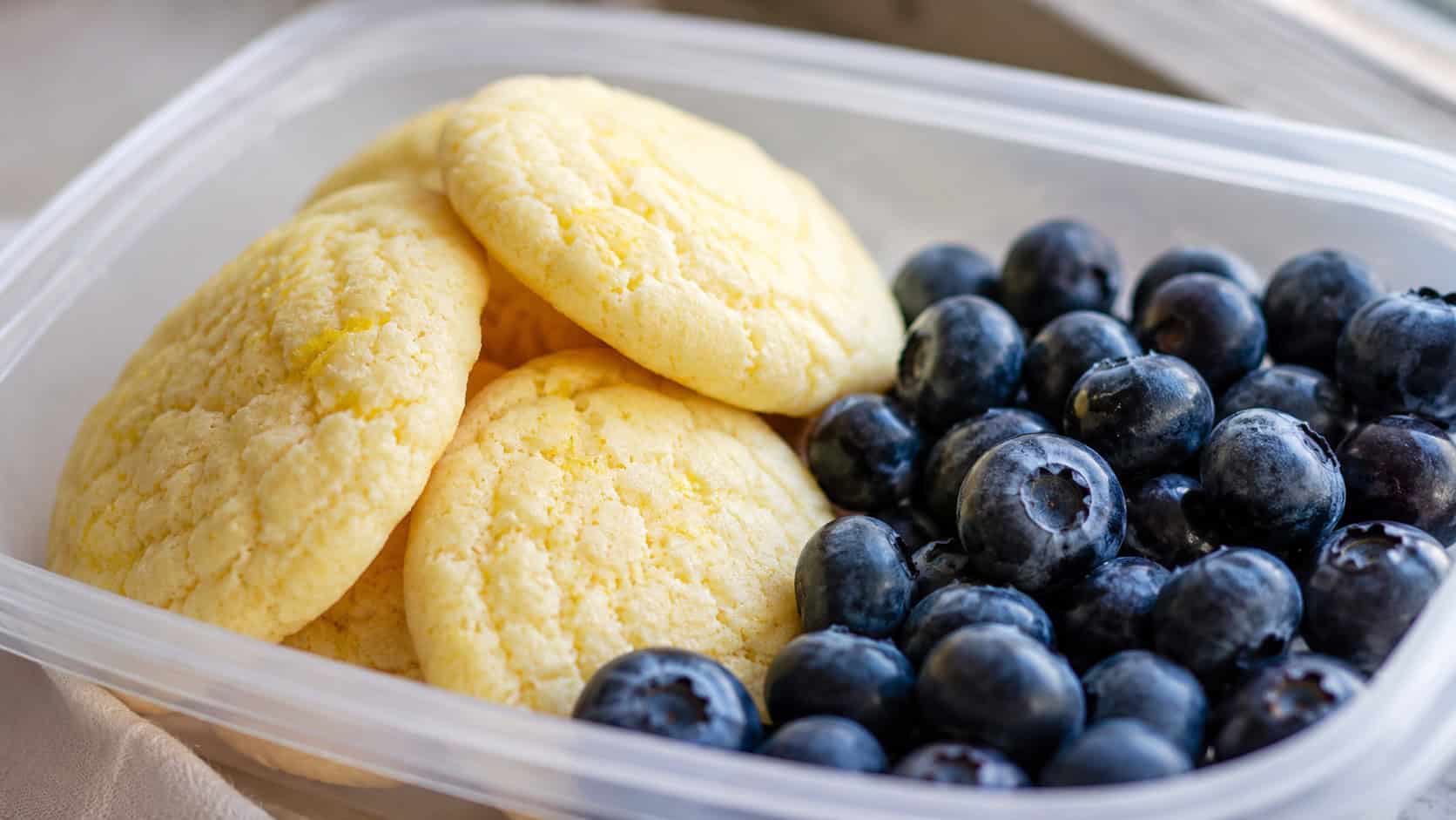 soft lemon cookies in a wide horizontal shot, pale yellow cookies beside a pile of fresh blueberries in a lunch container, bright natural light, shallow depth of field, landscape orientation
