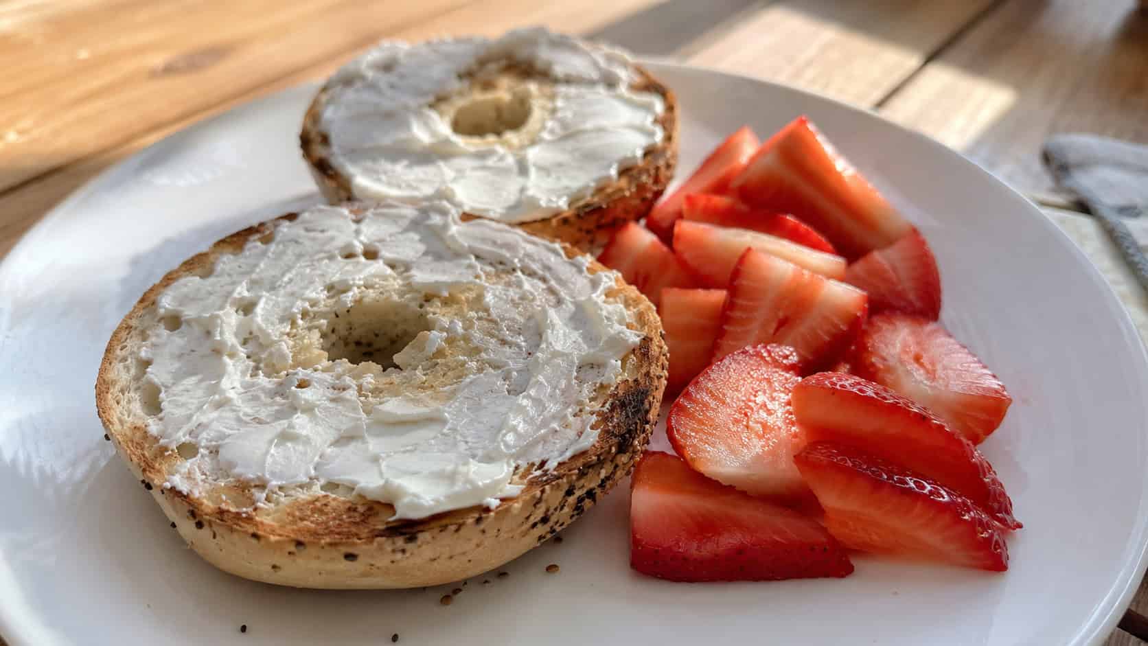 a toasted gluten-free bagel in a wide horizontal shot, halves spread with cream cheese beside sliced strawberries on a bright lunch plate, natural light, shallow depth of field, landscape orientation