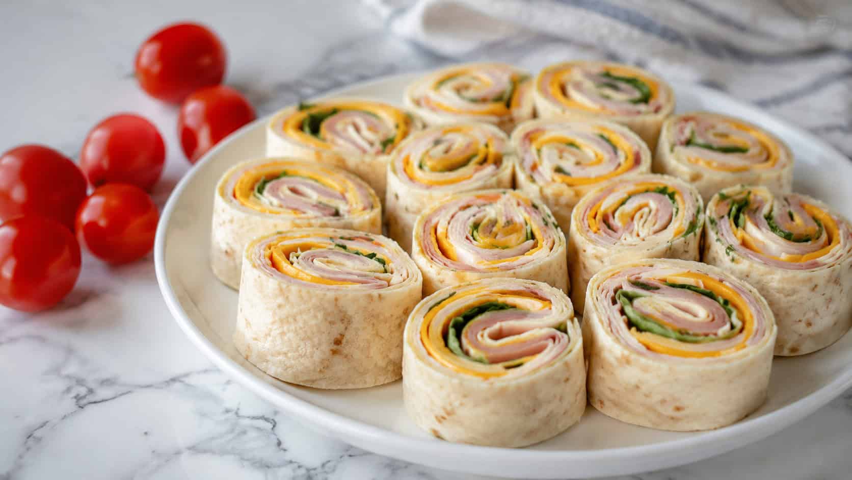 turkey and cheese roll-up pinwheels arranged cut-side up on a plate, shown in a wide horizontal shot on a marble counter with cherry tomatoes nearby, natural light, shallow depth of field, landscape orientation