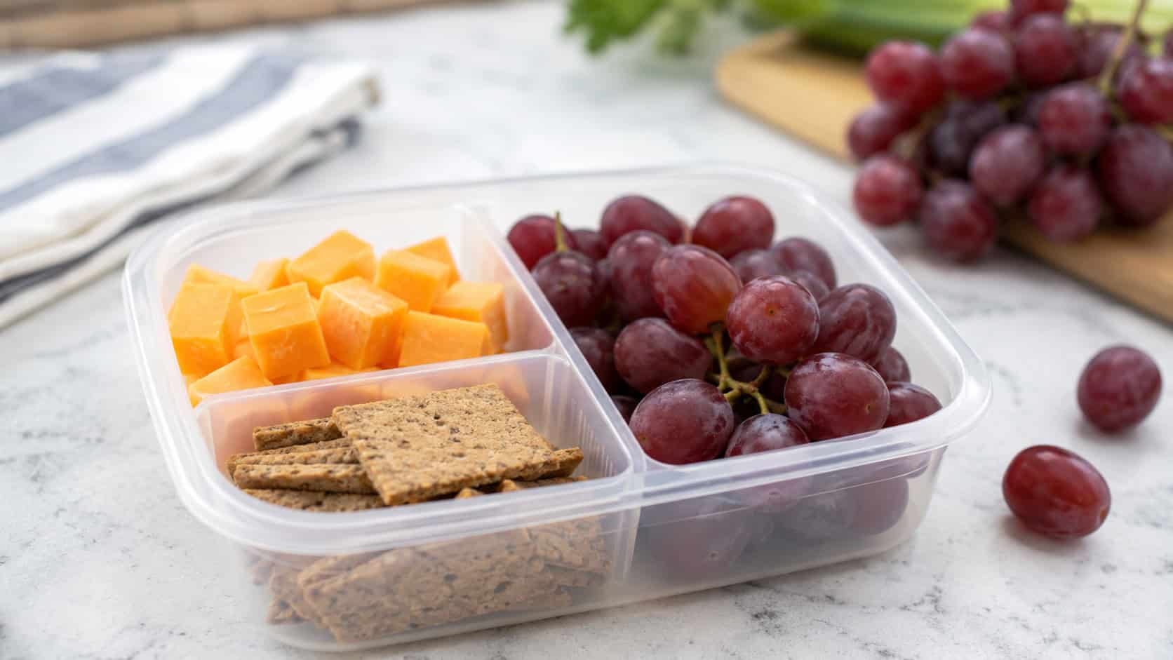 a snack lunch with cheddar cubes, whole grain crackers, and fresh grapes in a divided container, shown in a wide horizontal shot on a bright counter with extra grapes nearby, natural light, shallow depth of field, landscape orientation