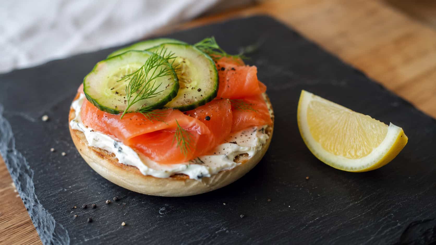 a mini bagel topped with cream cheese, smoked salmon, and cucumber, shown in a wide horizontal shot on a slate board with a lemon wedge nearby, natural light, shallow depth of field, landscape orientation