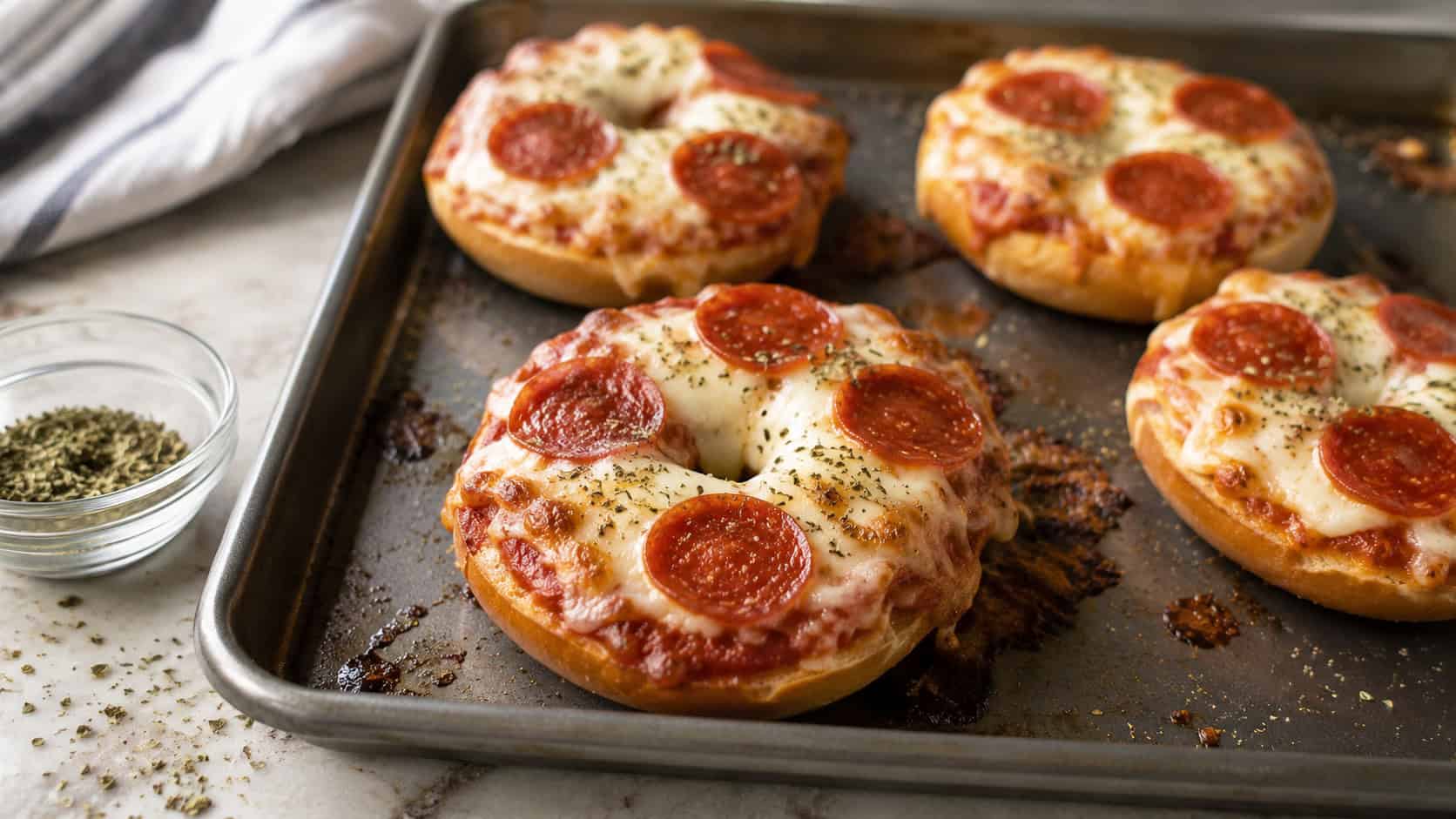 homemade pizza bagels with bubbly melted cheese and pepperoni on a baking sheet, shown in a wide horizontal shot on a kitchen counter with a sprinkle of oregano nearby, natural light, shallow depth of field, landscape orientation