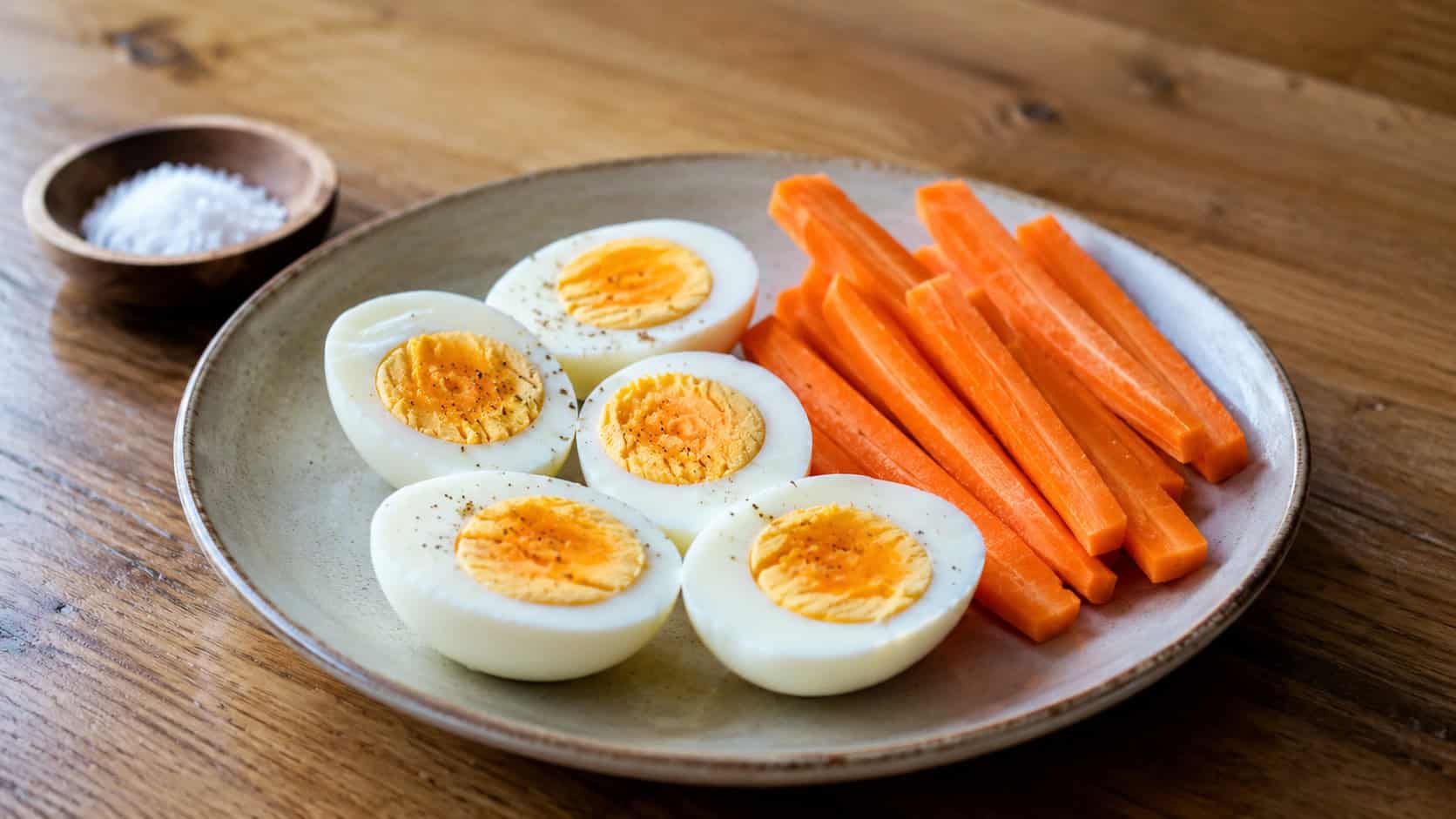 peeled hard-boiled eggs halved beside fresh carrot sticks on a plate, shown in a wide horizontal shot on a wooden table with a small dish of salt nearby, natural light, shallow depth of field, landscape orientation