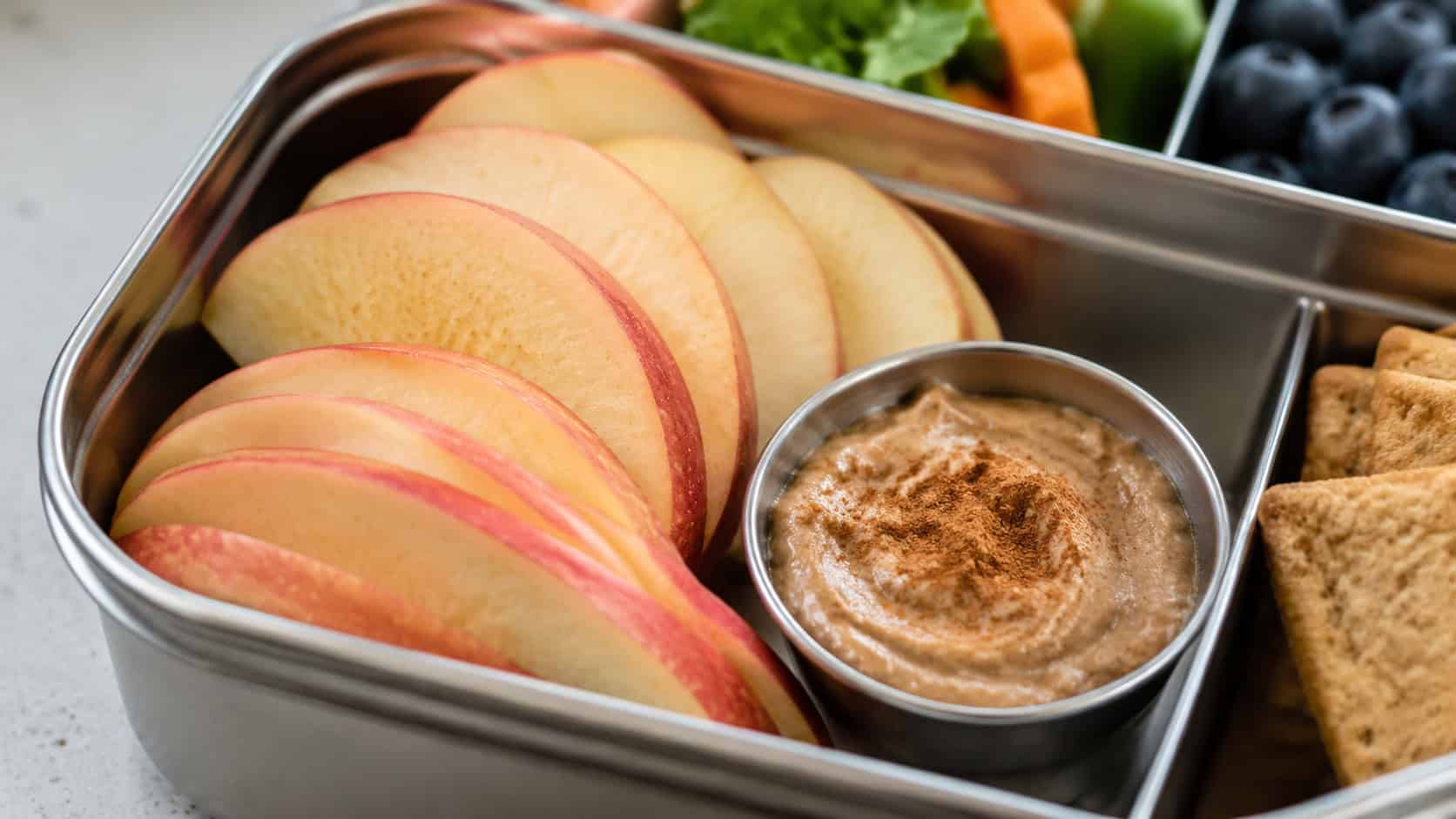 crisp apple slices fanned beside a small cup of creamy almond butter dusted with cinnamon, in a lunchbox compartment, in a wide horizontal shot, soft natural light, shallow depth of field, landscape orientation