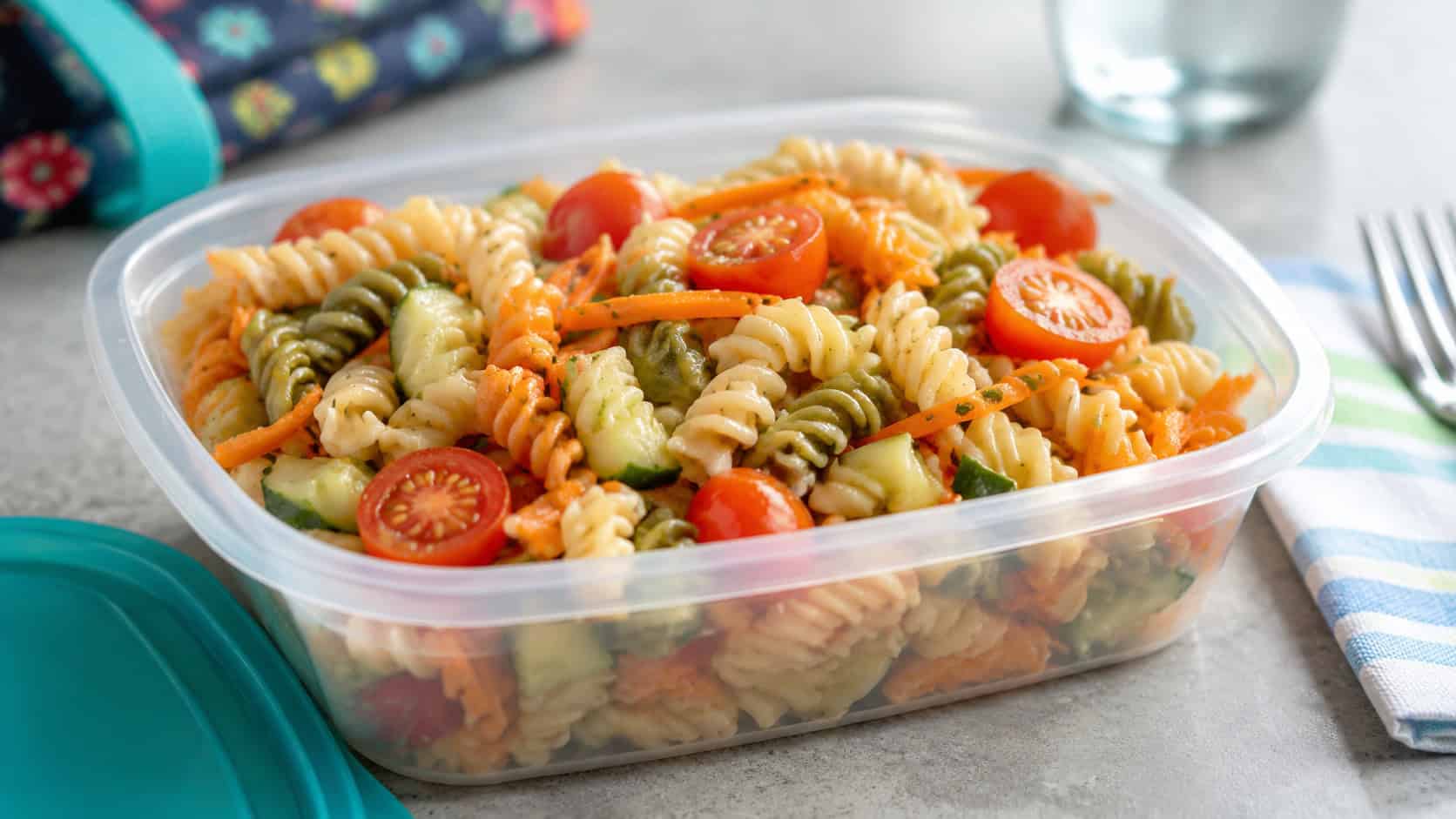 colorful veggie pasta salad with cherry tomatoes, cucumber, and shredded carrot in a small lunch container, in a wide horizontal shot, bright natural light, shallow depth of field, landscape orientation