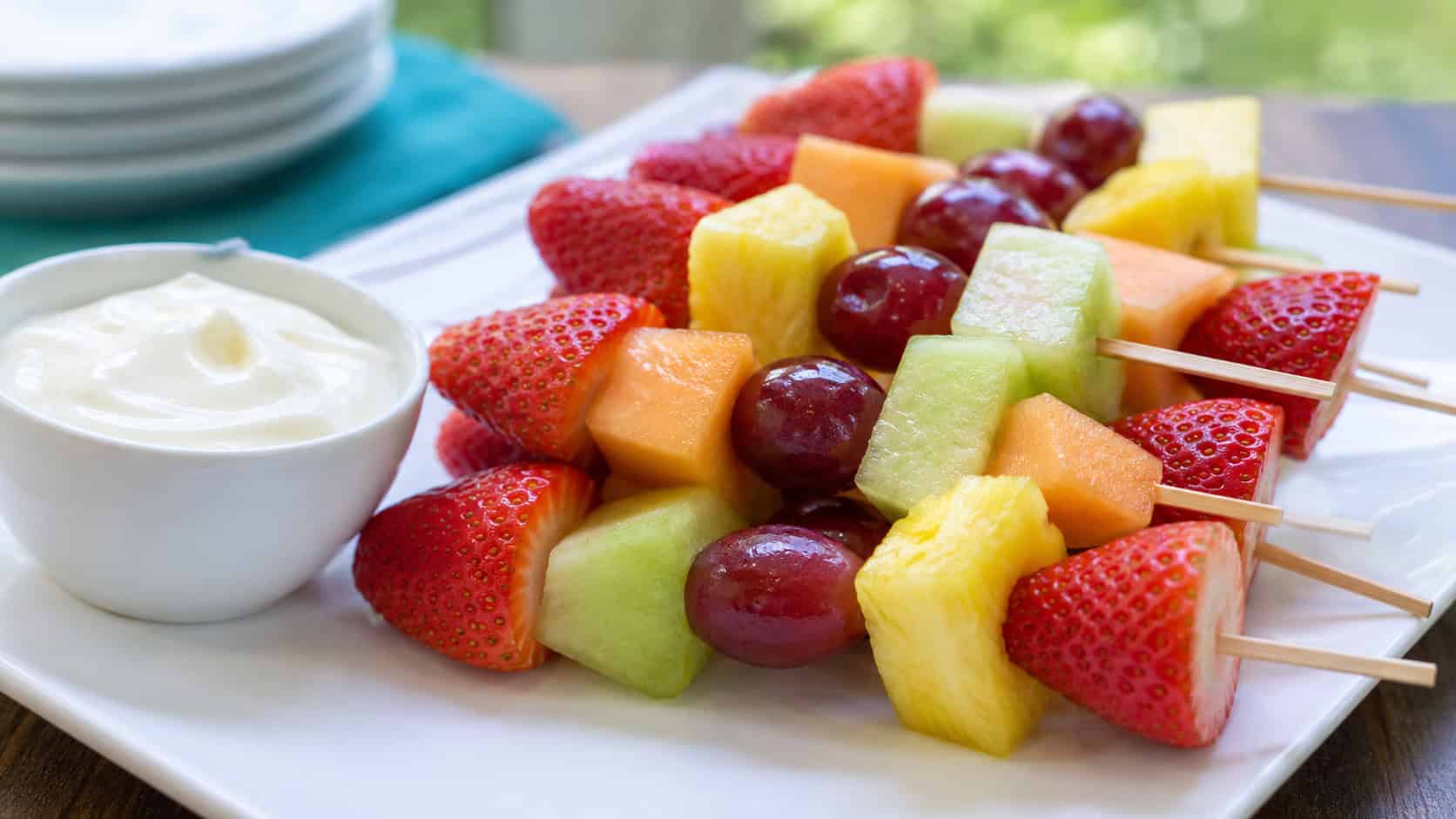 colorful fruit kabobs with strawberries, grapes, pineapple, and melon beside a small cup of creamy yogurt dip, on a bright plate, in a wide horizontal shot, natural daylight, shallow depth of field, landscape orientation