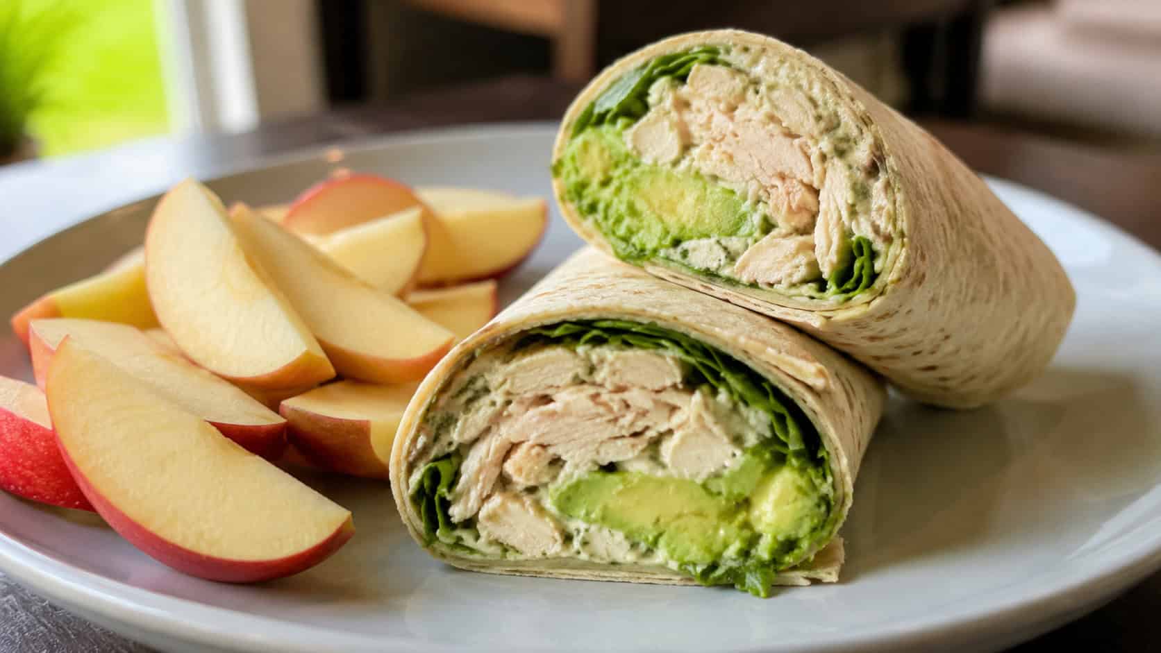 a chicken and avocado wrap sliced in half to show the green avocado and shredded chicken filling, on a plate beside apple slices, in a wide horizontal shot, bright natural light, shallow depth of field, landscape orientation