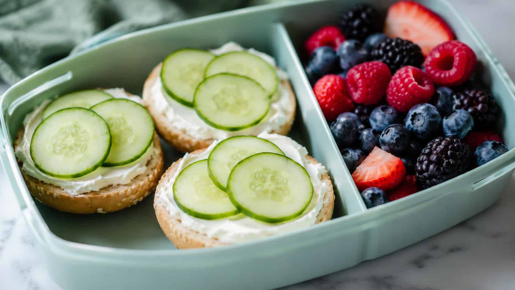 mini bagels spread with cream cheese and topped with cucumber rounds, arranged in a lunchbox with a handful of berries, in a wide horizontal shot, soft natural light, shallow depth of field, landscape orientation