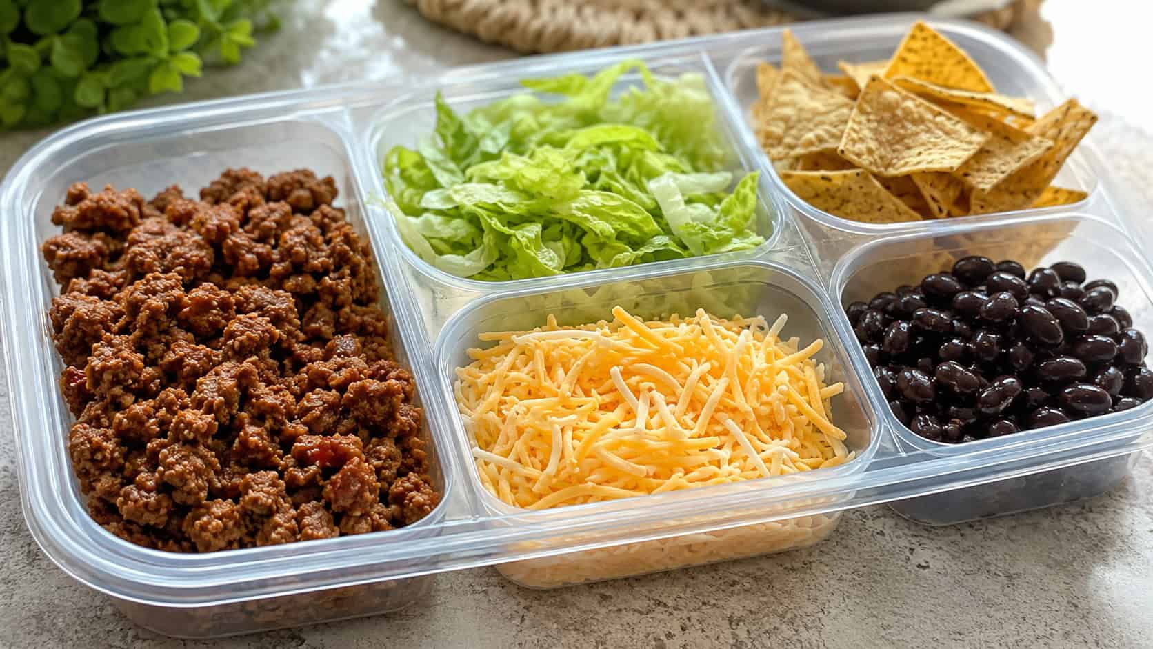a build-your-own taco salad lunchbox with seasoned ground turkey, shredded lettuce, cheese, black beans, and tortilla chips in separate compartments, in a wide horizontal shot, bright natural light, shallow depth of field, landscape orientation