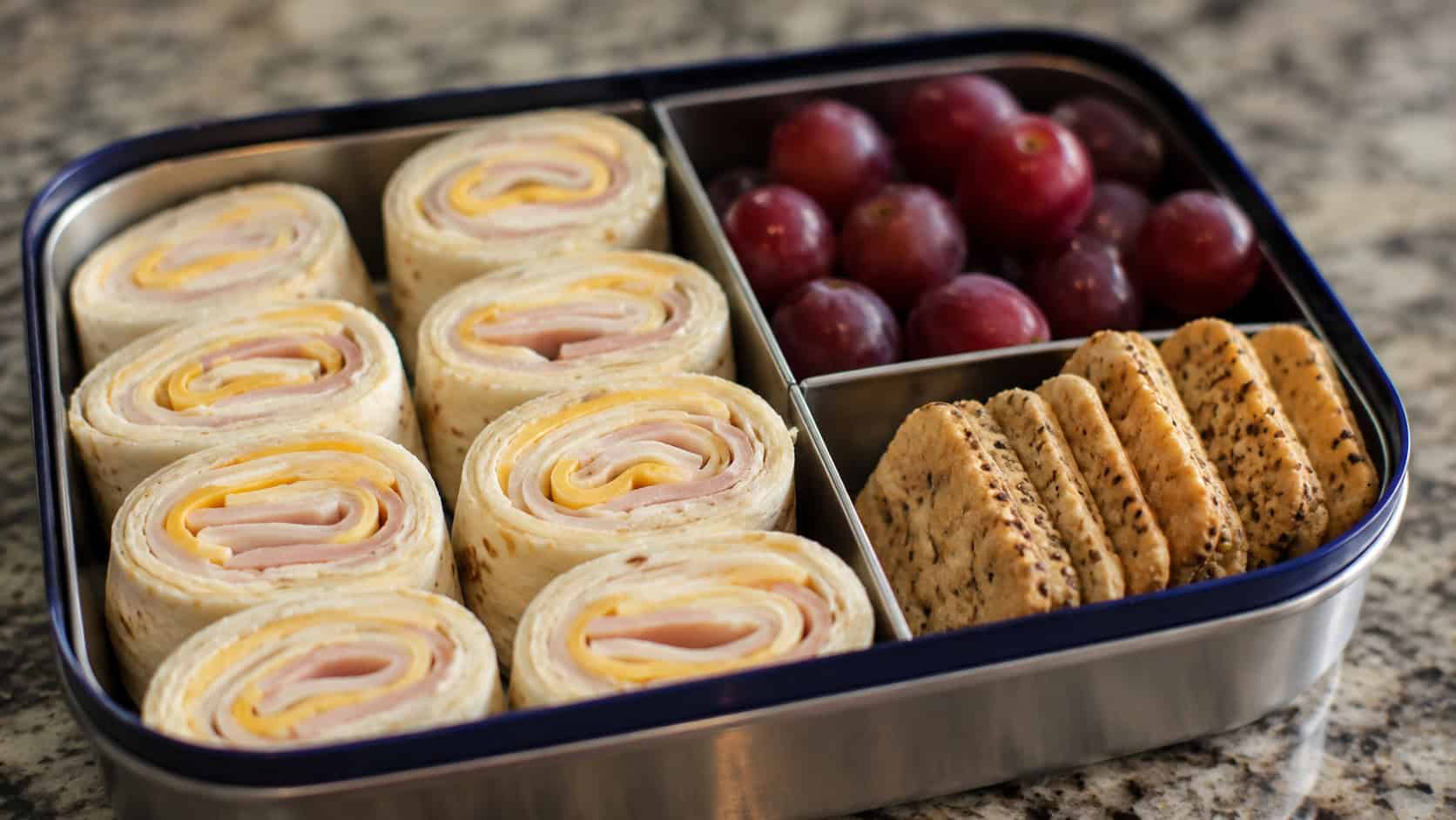 turkey and cheese pinwheel roll-ups in a wide horizontal shot, arranged cut-side up in a bento lunchbox beside grapes and crackers on a kitchen counter, natural light, shallow depth of field, landscape orientation