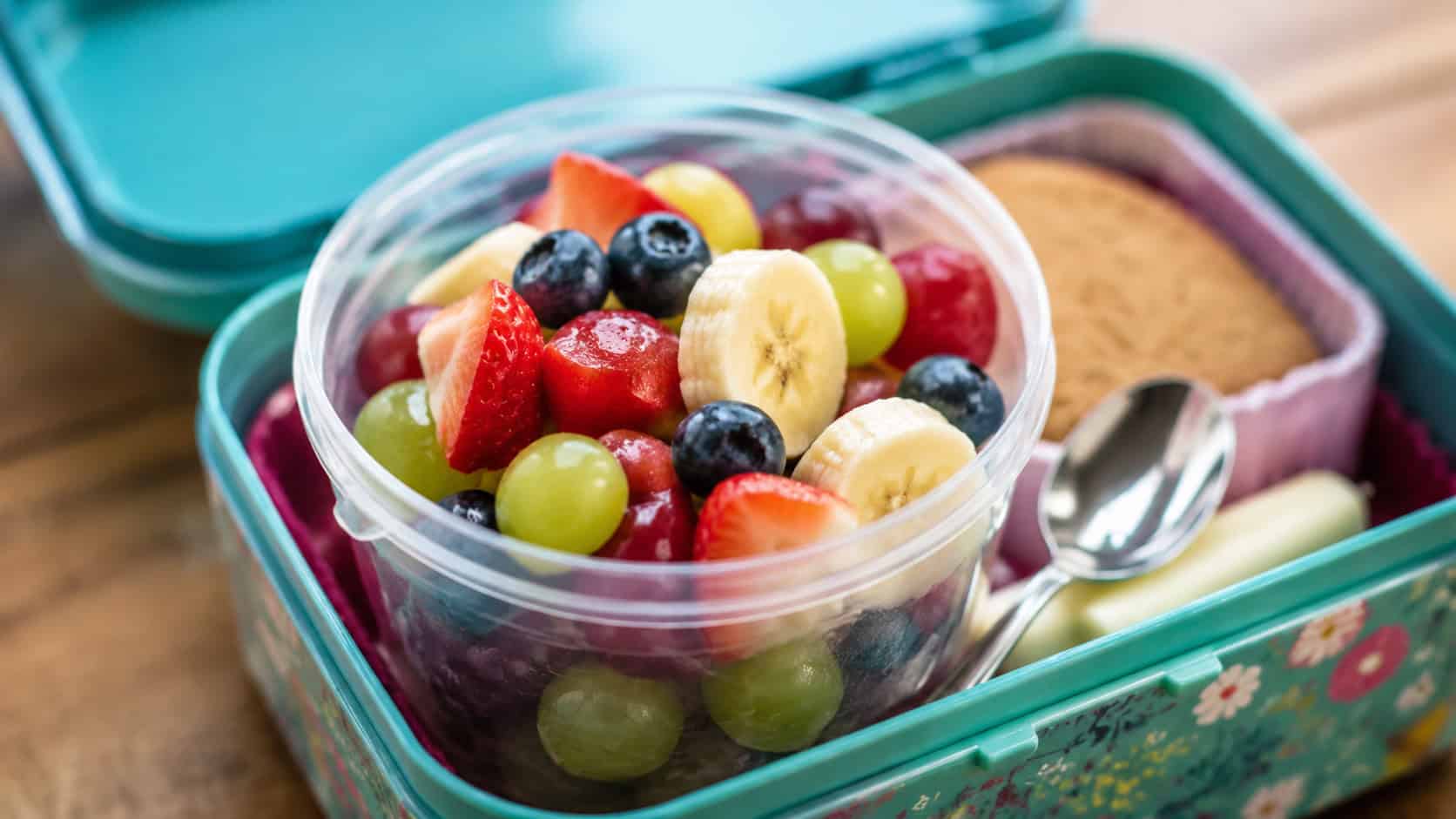a colorful fruit salad cup full of strawberries, grapes, banana, and blueberries in a wide horizontal shot, set in an open lunchbox with a small spoon, natural light, shallow depth of field, landscape orientation