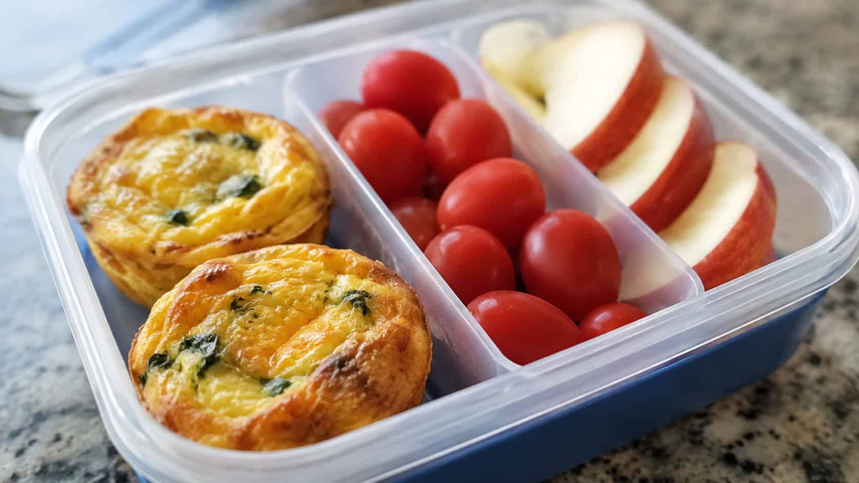 golden mini quiches in a wide horizontal shot, two tucked into a lunchbox compartment next to cherry tomatoes and apple slices, natural light, shallow depth of field, landscape orientation