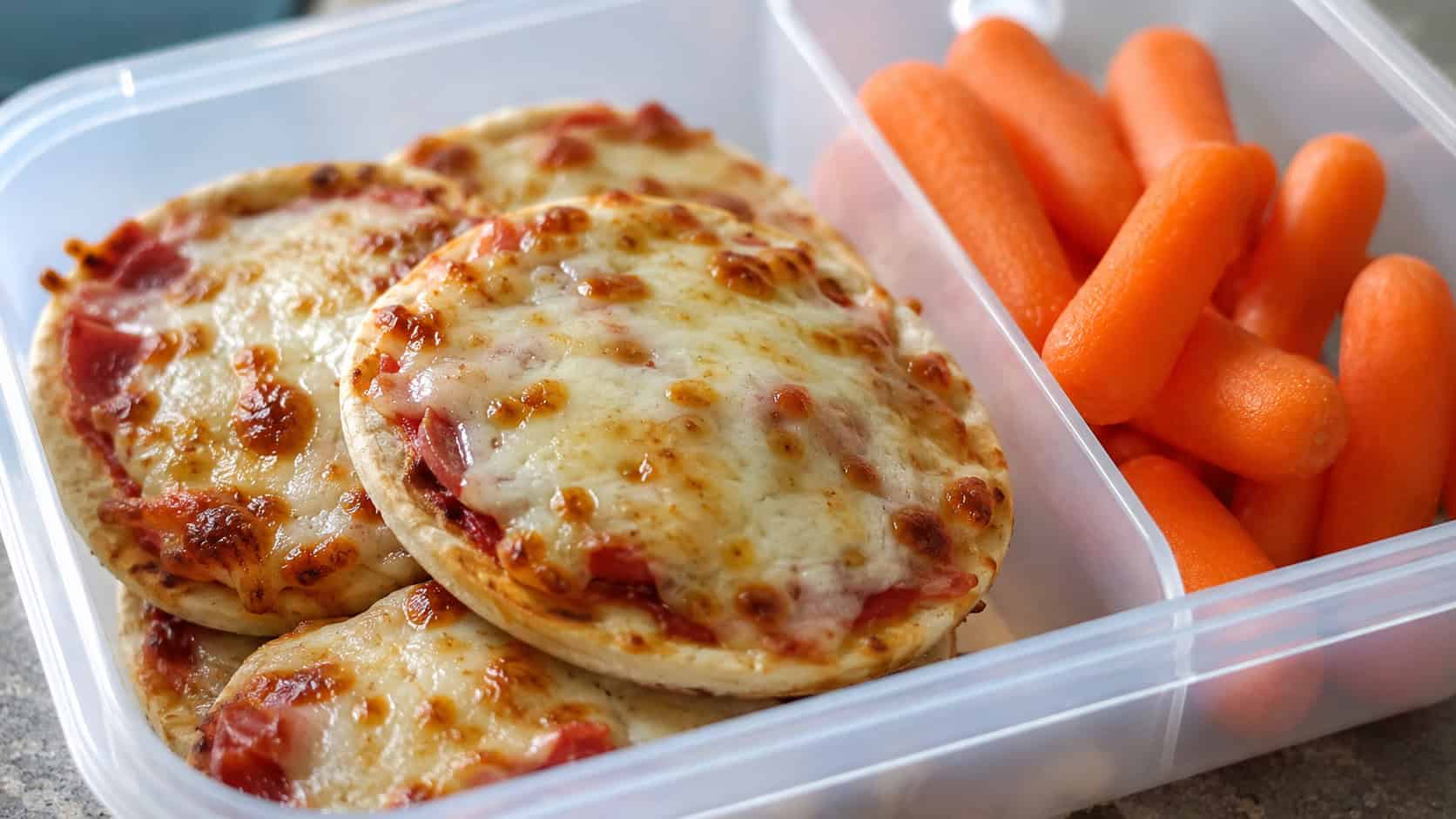 mini pita pizzas with melted bubbly cheese in a wide horizontal shot, arranged in a lunchbox compartment next to baby carrots, natural light, shallow depth of field, landscape orientation
