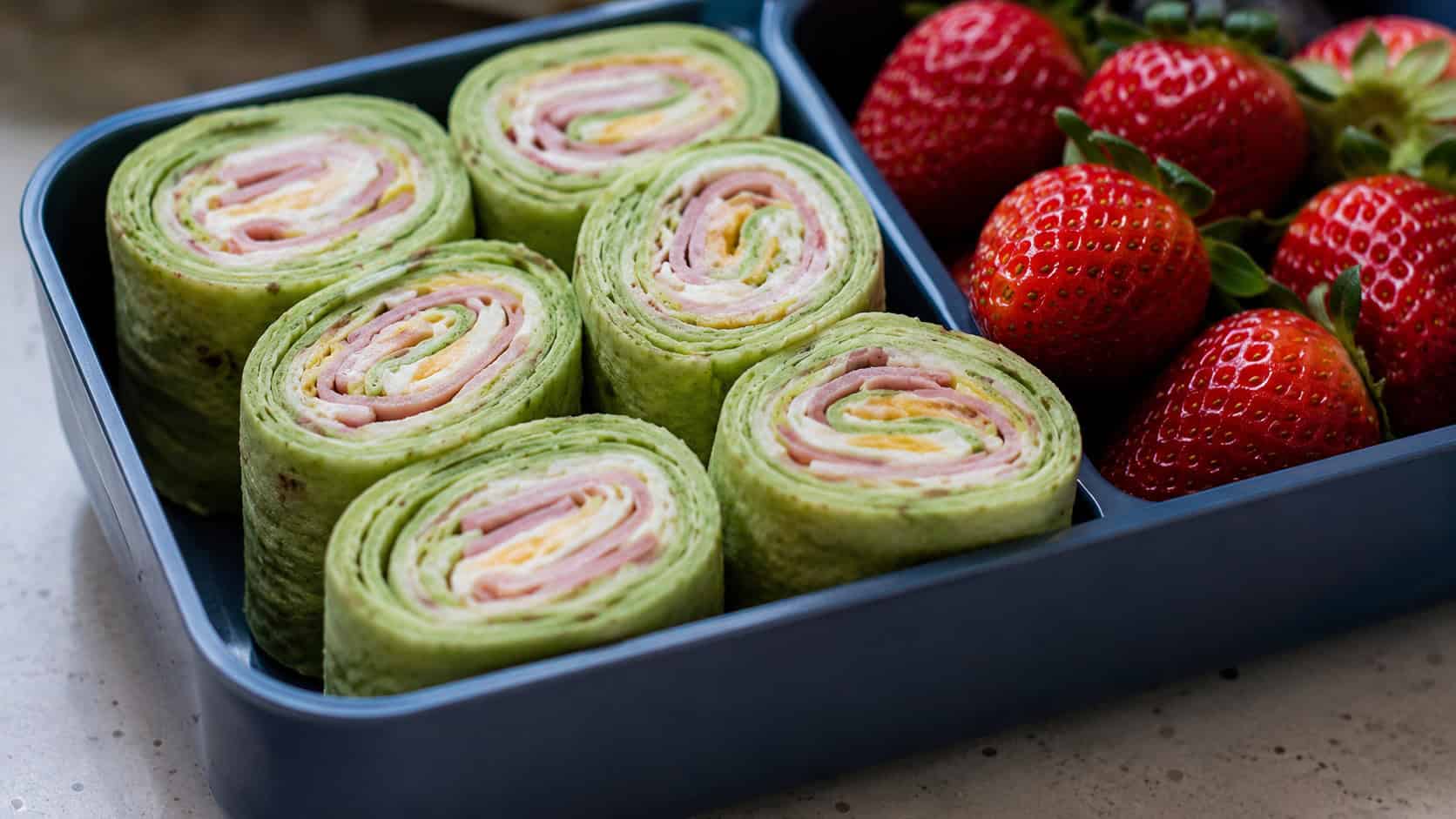 green-tinted tortilla pinwheels with ham and cheese in a wide horizontal shot, lined up cut-side up in a bento box beside strawberries, natural light, shallow depth of field, landscape orientation