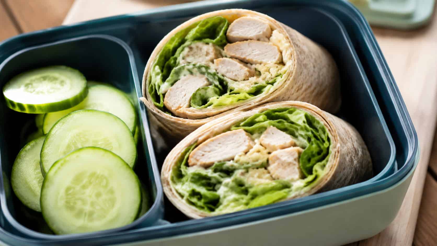 a chicken Caesar wrap sliced in half in a wide horizontal shot, showing the layered filling, tucked into a lunchbox beside cucumber slices, natural light, shallow depth of field, landscape orientation