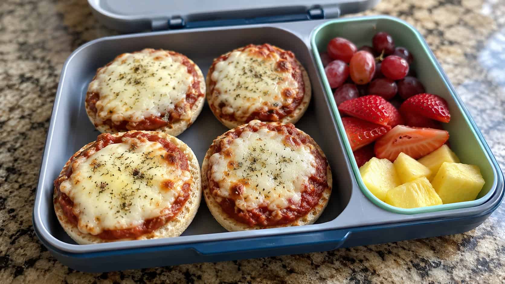 four mini English muffin pizzas with melted cheese in a wide horizontal lunchbox, with fruit in the next compartment, set on a counter, bright natural light, shallow depth of field, landscape orientation