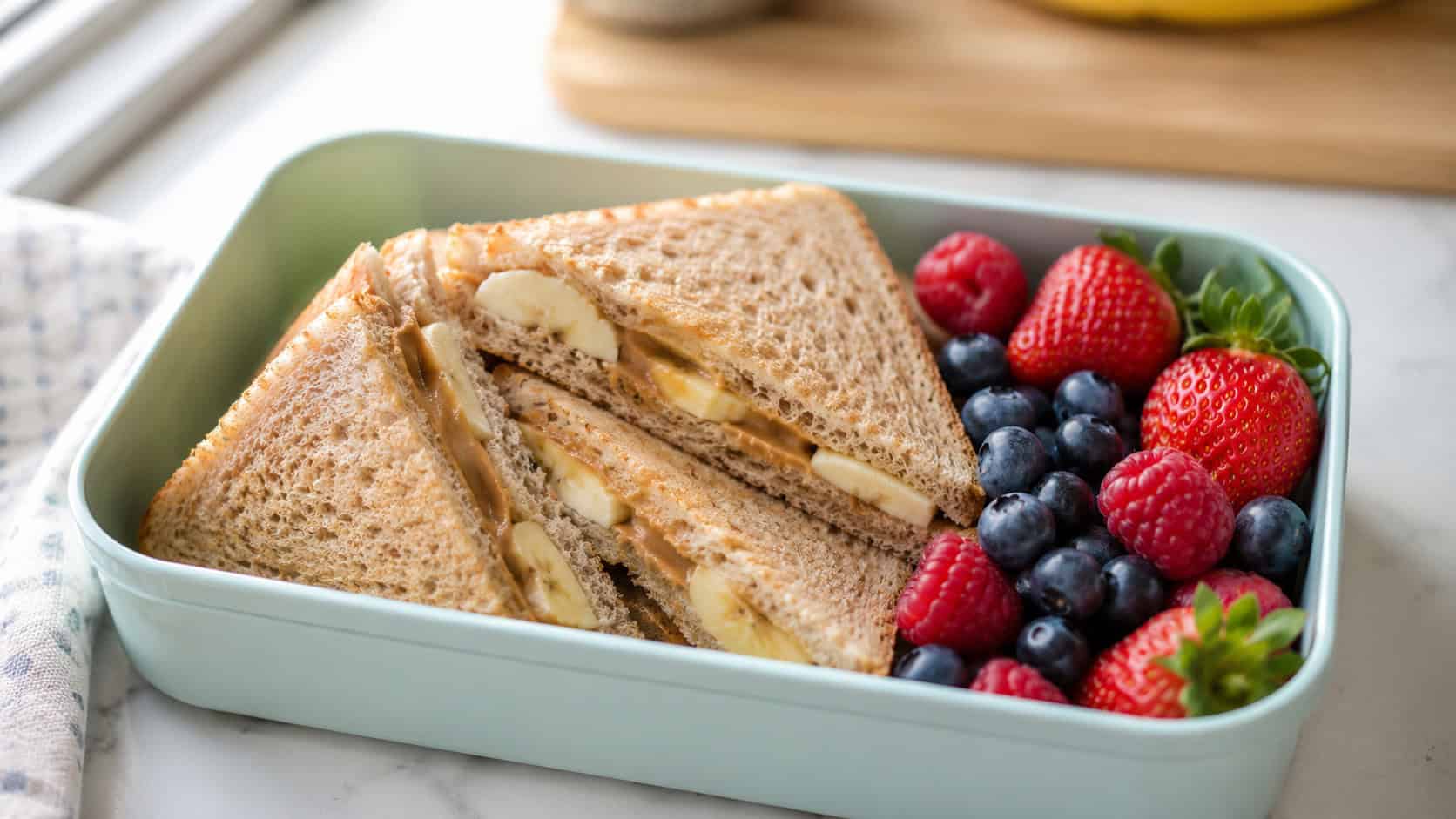 a peanut butter and banana sandwich cut into triangles in a wide horizontal lunchbox, with berries on the side, set on a bright kitchen table, natural light, shallow depth of field, landscape orientation
