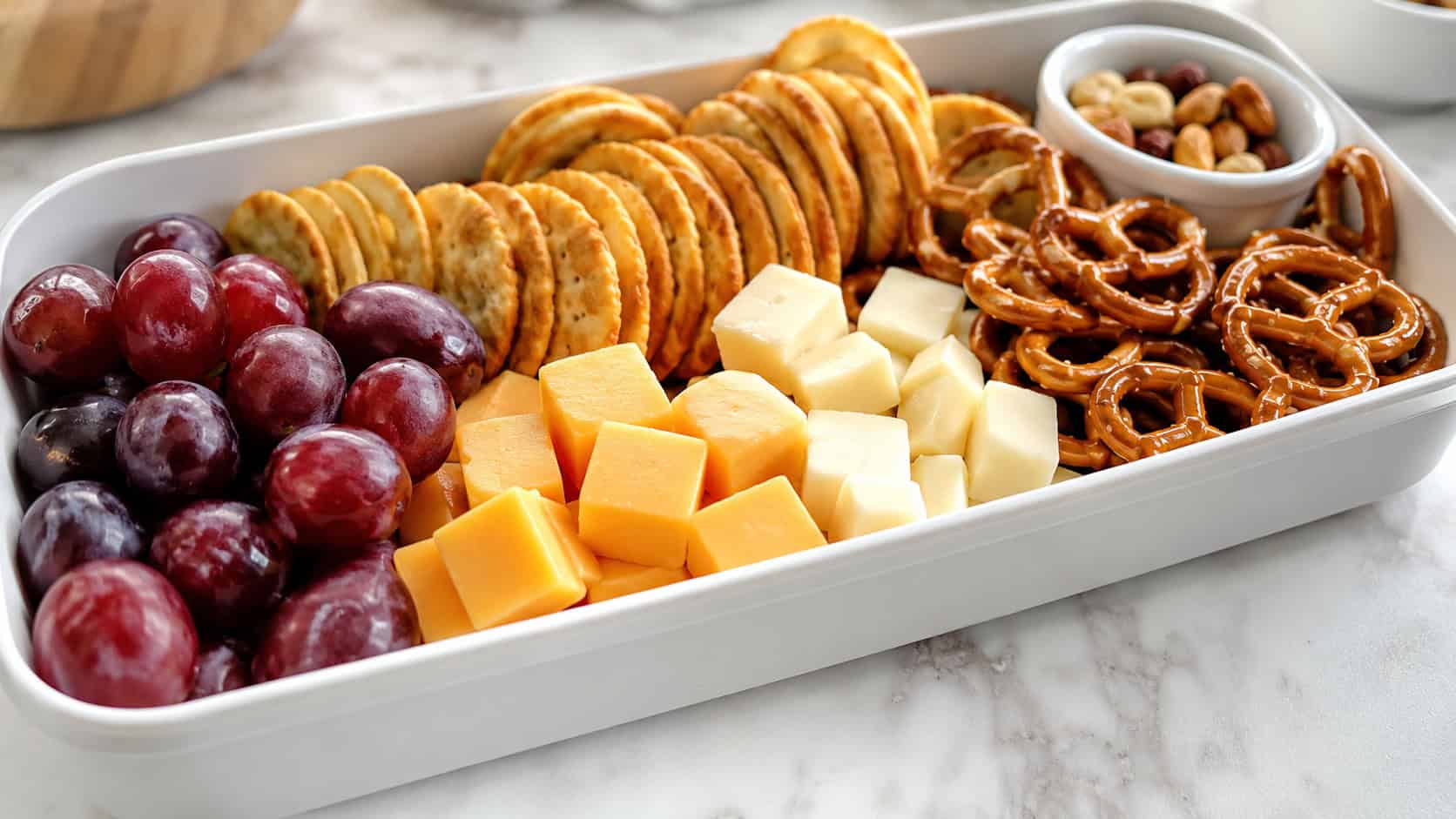 a colorful snack board with cheese cubes, crackers, grapes, and pretzels in a wide horizontal lunchbox, set on a bright counter, natural light, shallow depth of field, landscape orientation