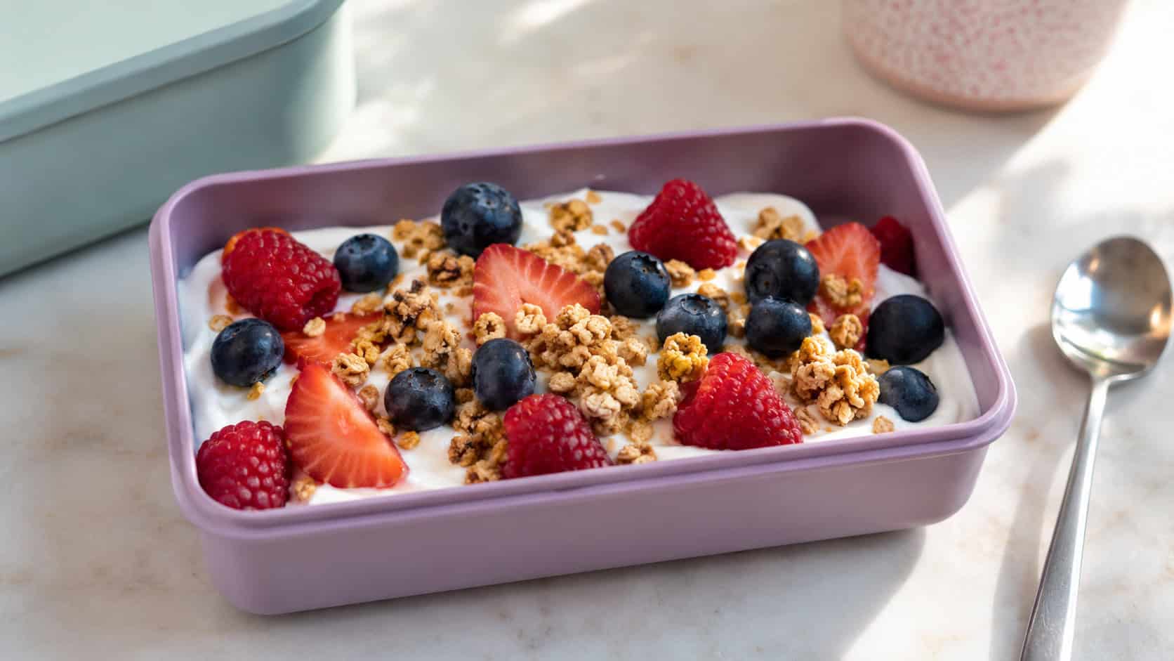 a cup of creamy yogurt topped with granola and fresh berries in a wide horizontal lunchbox, with a small spoon beside it, set on a bright table, natural light, shallow depth of field, landscape orientation