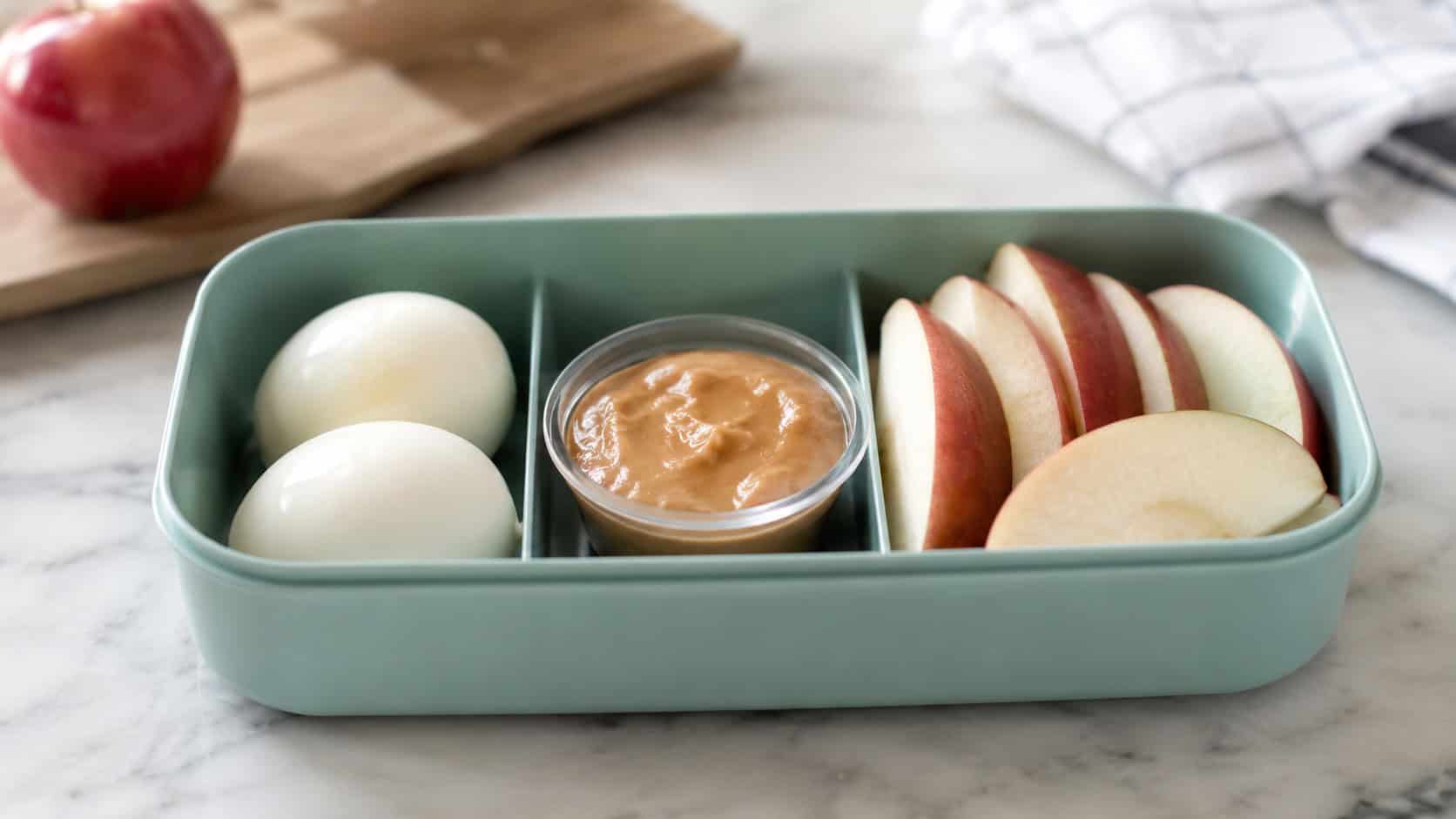 peeled hard-boiled eggs and crisp apple slices in a wide horizontal lunchbox, with a small container of dip, set on a bright kitchen counter, natural light, shallow depth of field, landscape orientation