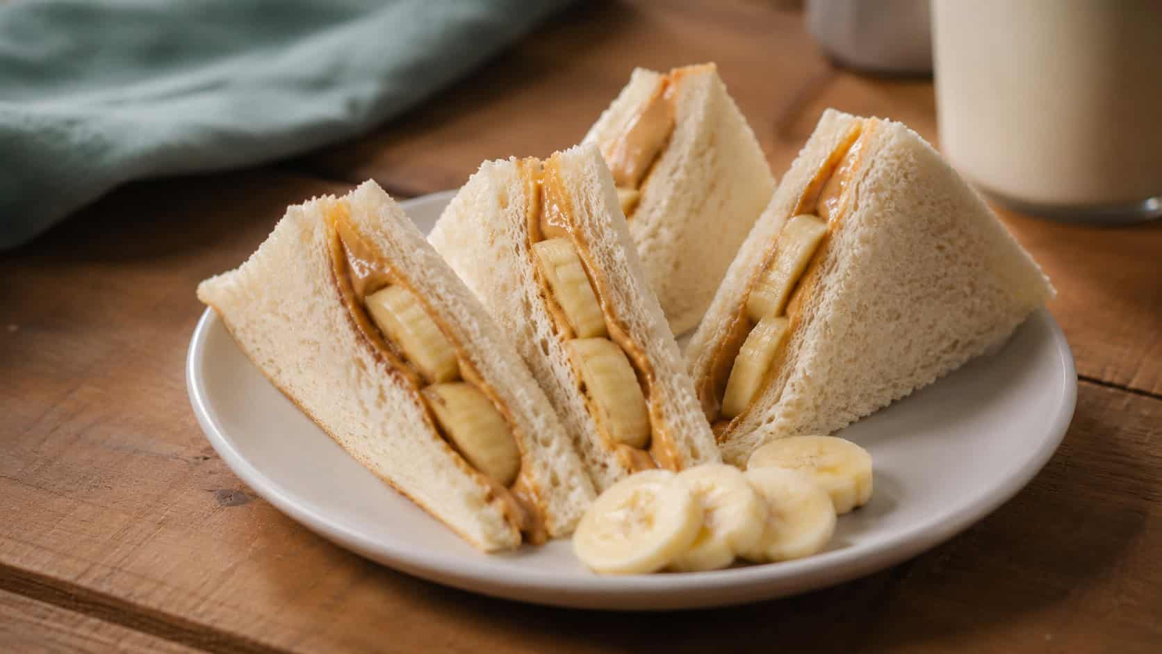 a peanut butter and banana sandwich cut into triangles and fanned out in a wide horizontal arrangement on a small plate, soft bread and banana slices peeking out, a few extra banana coins nearby, warm natural light, shallow depth of field, landscape orientation
