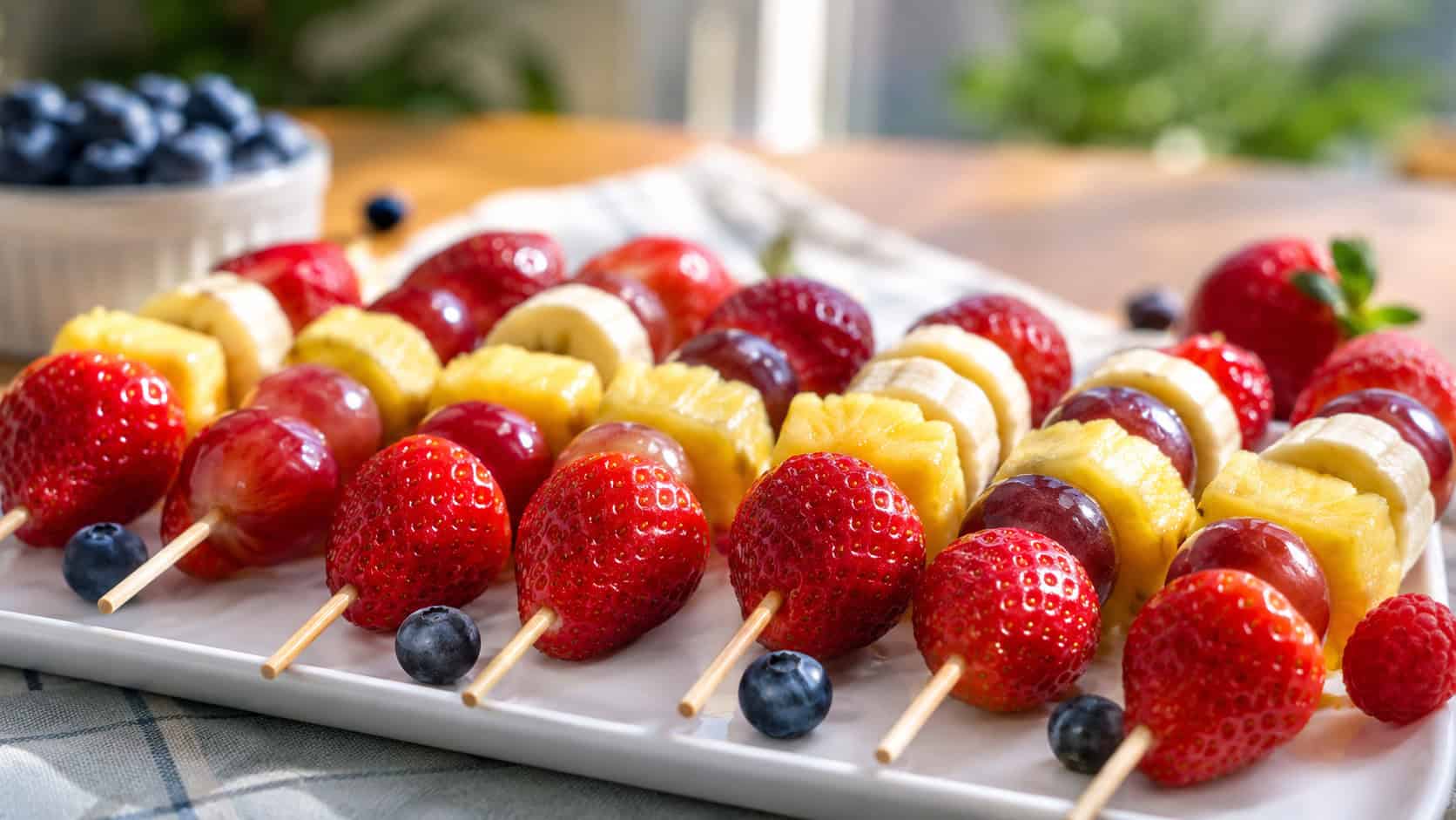 colorful fresh fruit kabobs lined up in a wide horizontal row on a white platter, strawberries grapes pineapple and banana glistening, a few loose berries scattered around, sunny kitchen table, natural light, shallow depth of field, landscape orientation