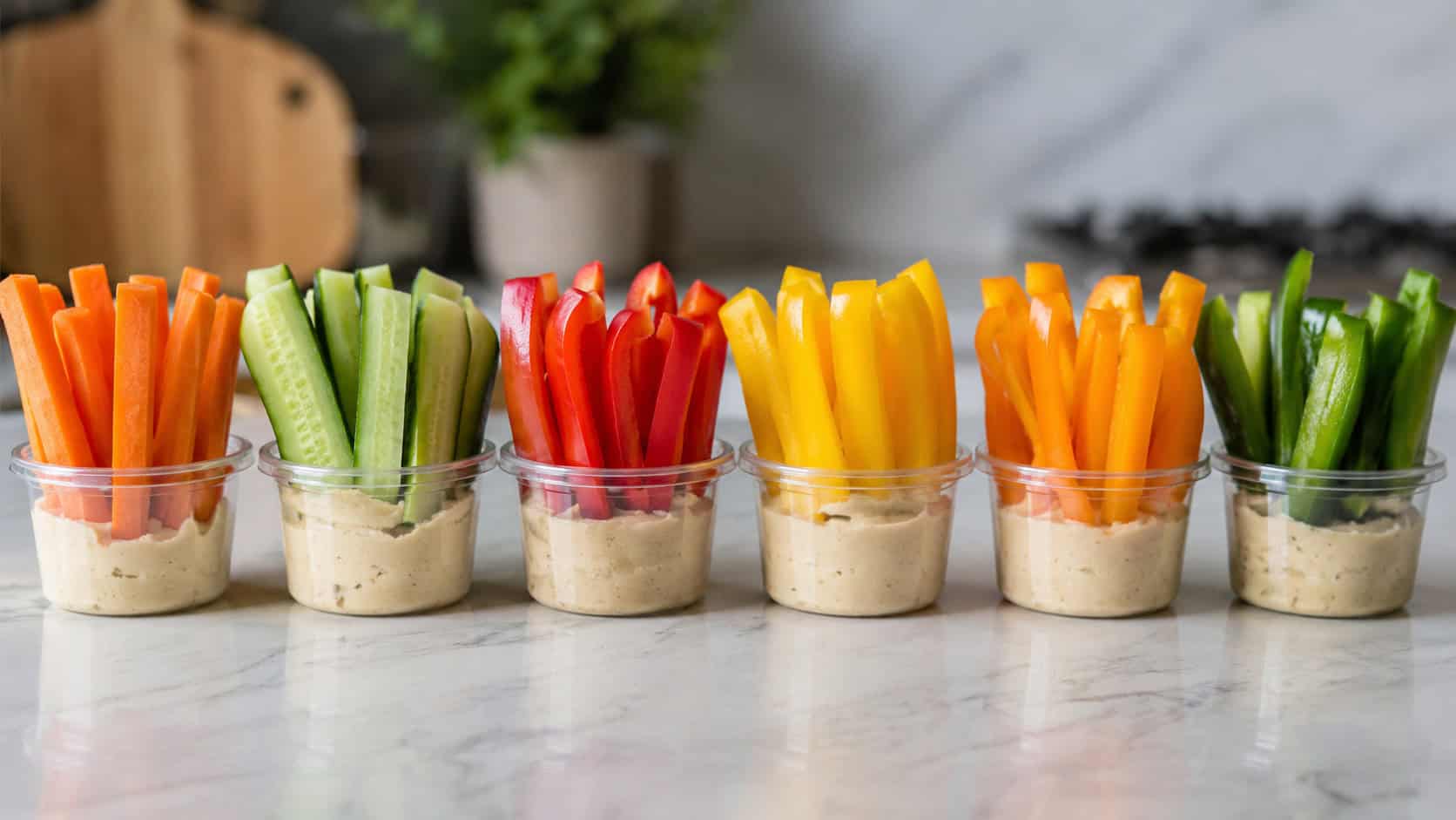 small cups filled with hummus and upright colorful veggie sticks arranged in a wide horizontal line on a marble counter, carrots cucumbers and bell peppers vivid and fresh, soft natural light, shallow depth of field, landscape orientation